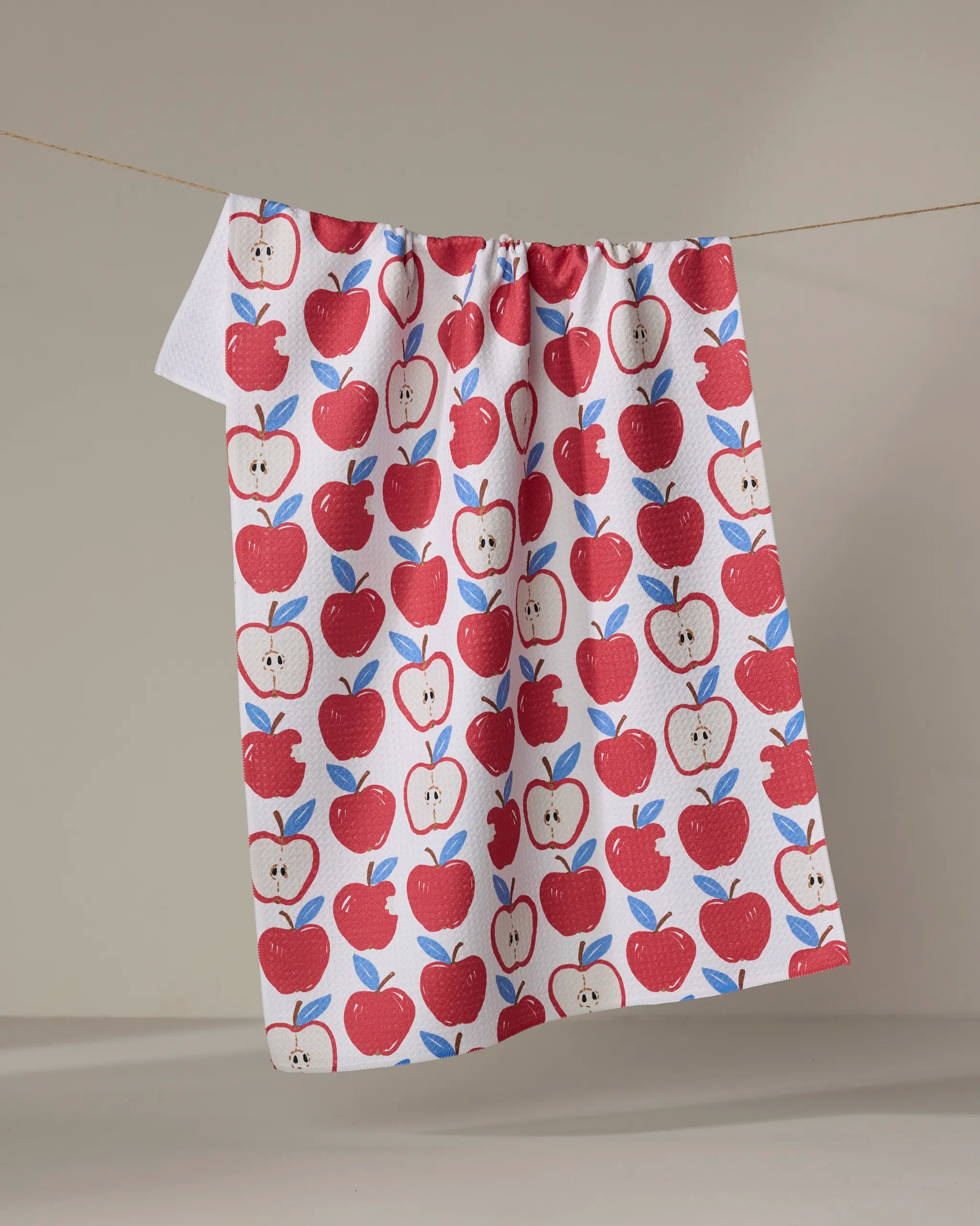 The Apple A Day tea towel by Hufton Studio features red apples and green leaves on white, hanging against a neutral background. Made from recycled materials, this absorbent towel brings eco-friendly charm to your kitchen.