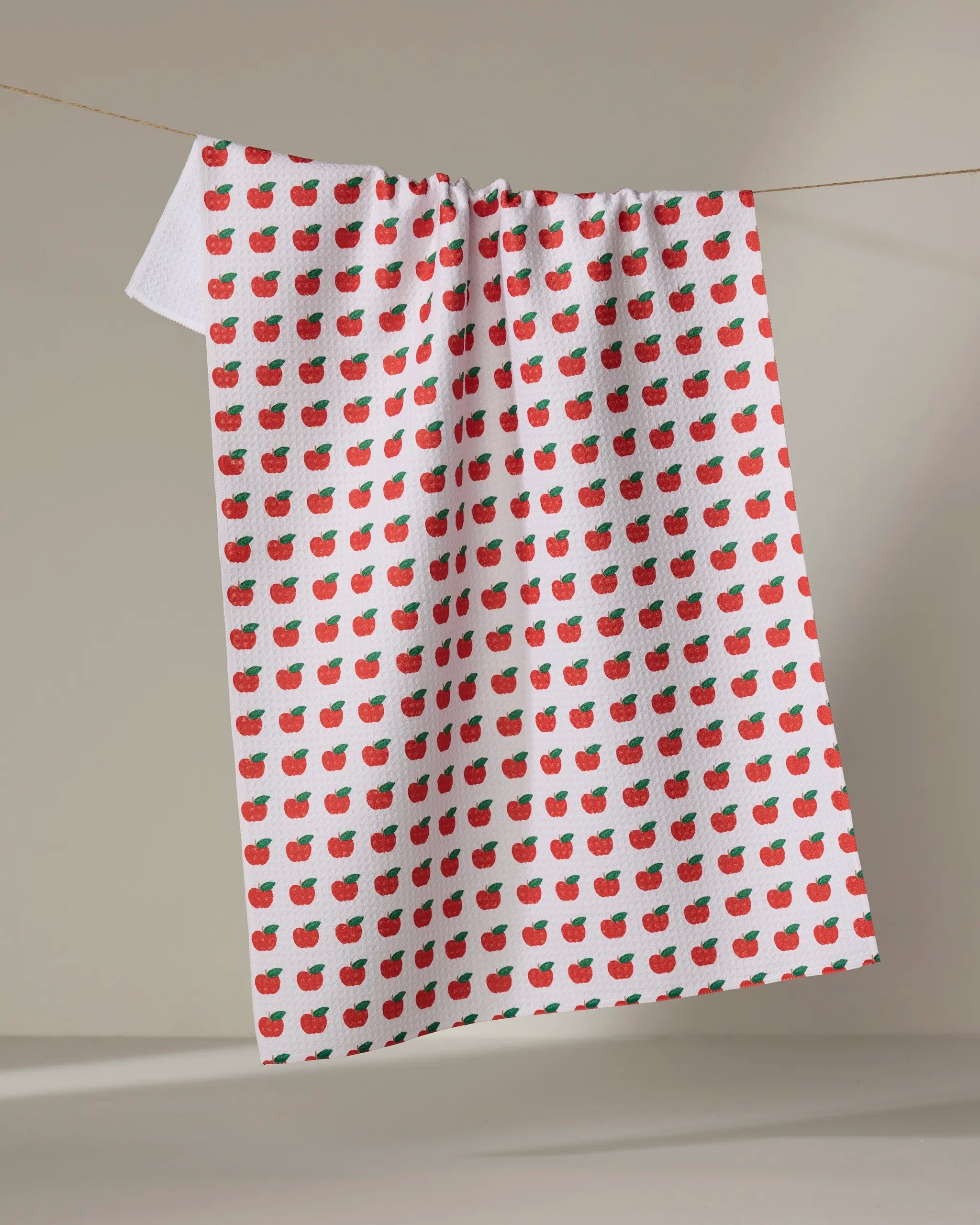 The Apple Grove tea towel by Brittany Jepsen features a white textured design with small red apples and green leaves, made from recycled materials. Quick-drying and softly lit on a neutral background, it hangs gracefully on a line.