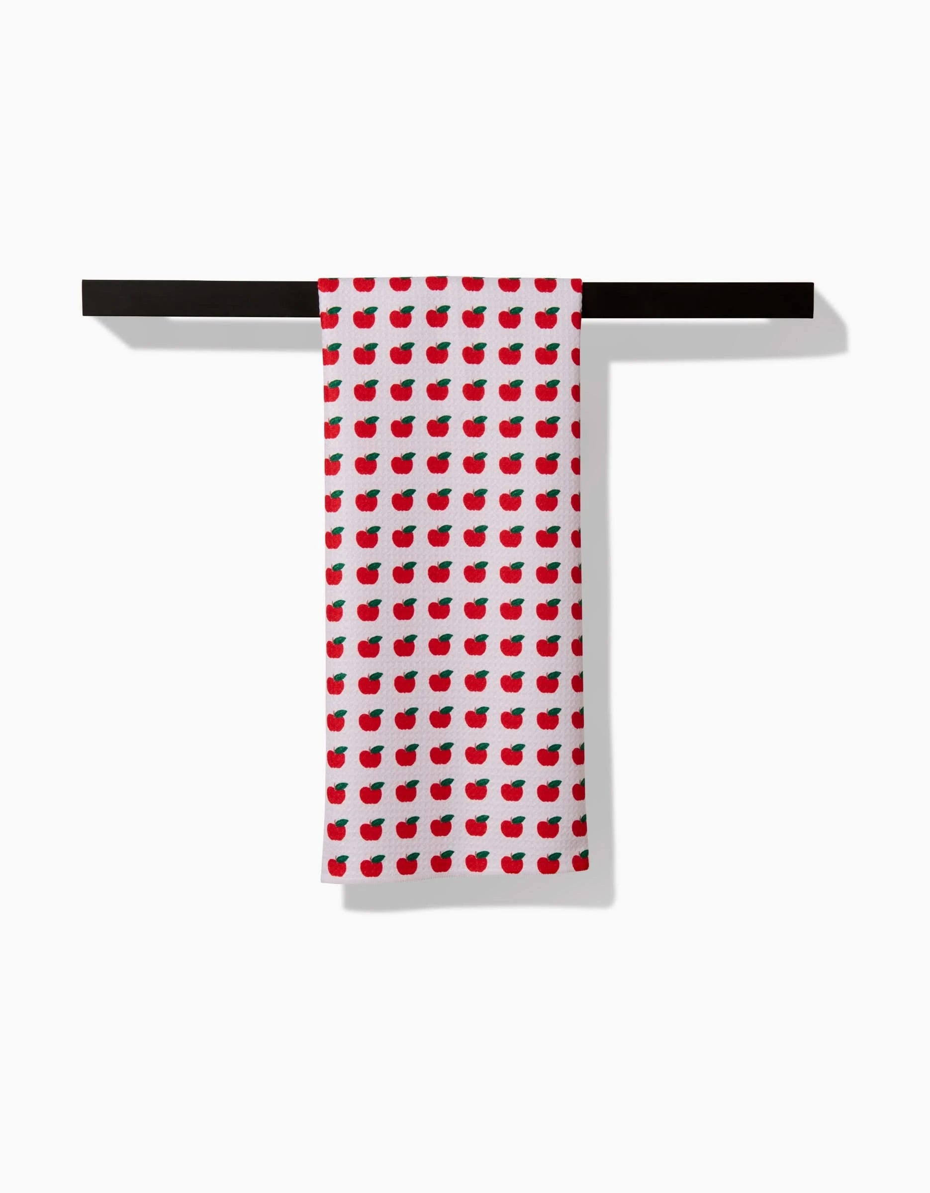 The Apple Grove tea towel by Brittany Jepsen, made from Geoweave fabric, features a repeating red apple and green leaf pattern and is displayed draped over a black horizontal bar on a plain white background.