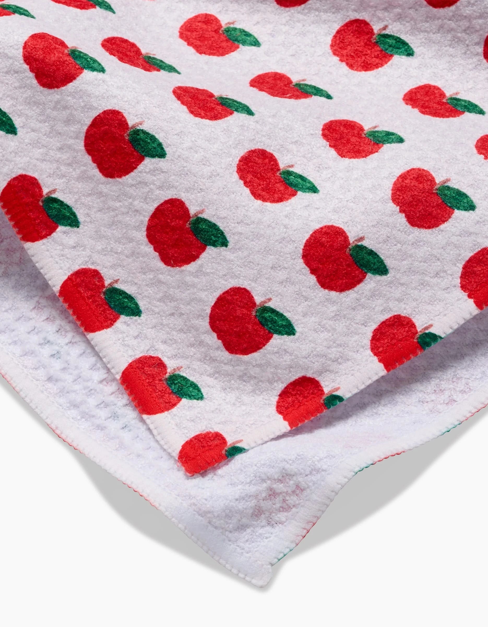 A close-up of the Apple Grove tea towel by Brittany Jepsen, showing bright red apples with green leaves on white Geoweave fabric, with visible textured edges and slight overlap.