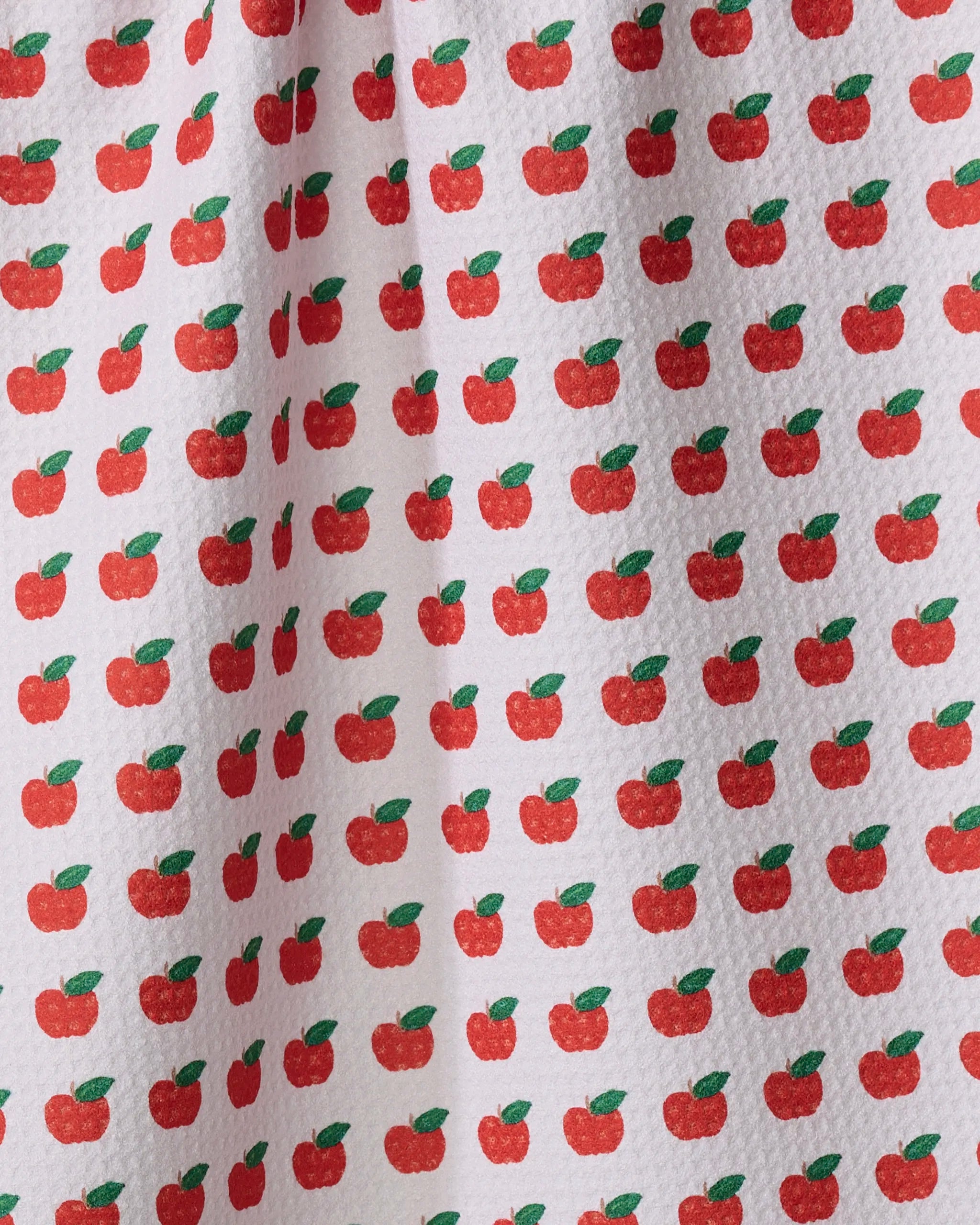 Close-up of the Brittany Jepsen "Apple Grove" tea towel, crafted from recycled materials and adorned with a neat pattern of small red apples with green leaves across its textured white surface.