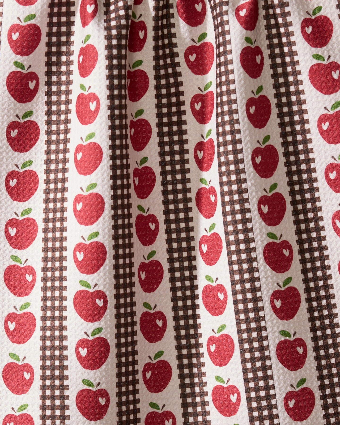 The Geometry “Apple Picking Day” tea towel features red apples with white highlights in vertical rows, alternating with brown plaid stripes on a textured white background for a charming, vintage country kitchen look.