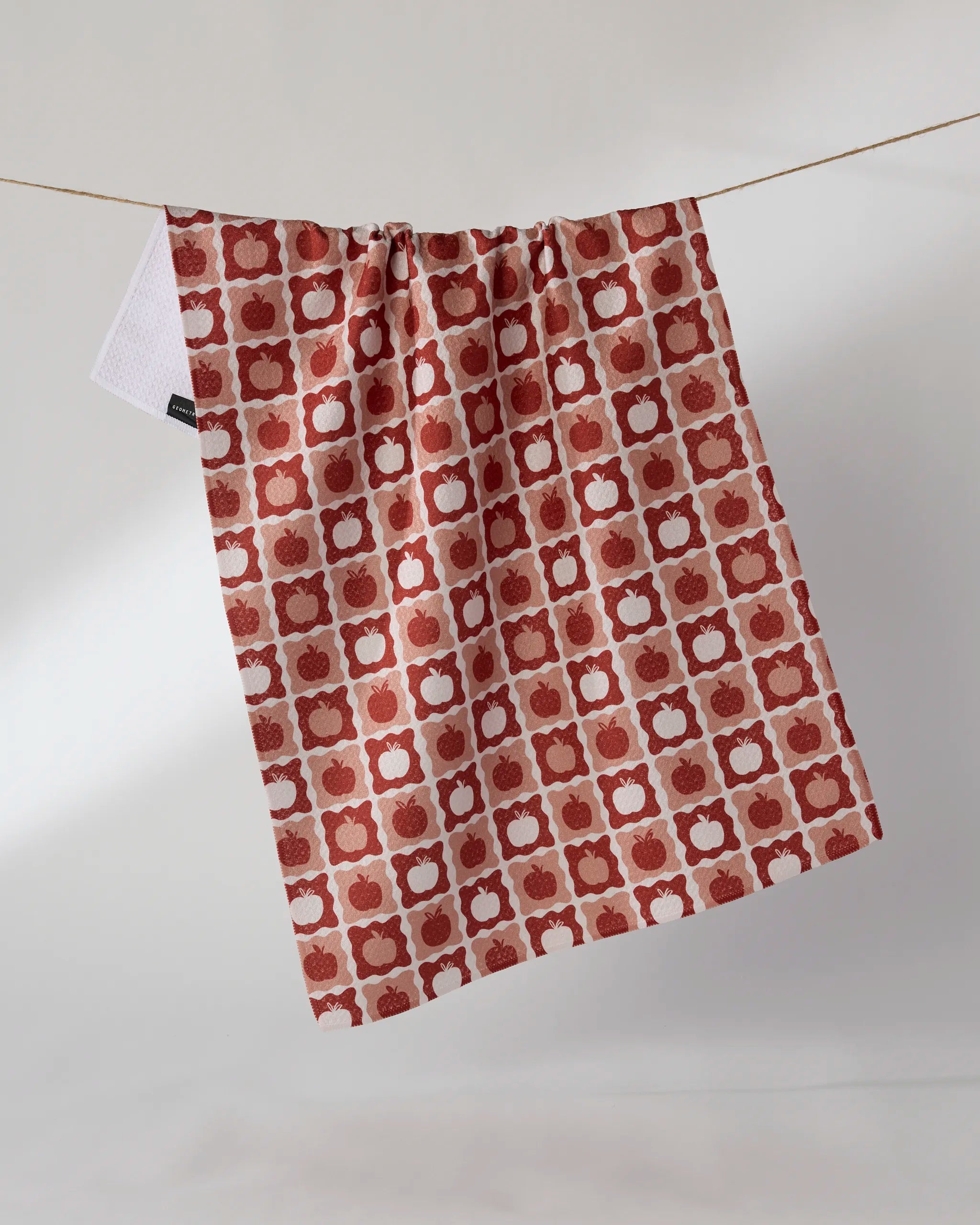 The Geometry Apple Quilt, a recycled kitchen towel featuring a red and white geometric apple pattern, hangs on a string against a plain white background.