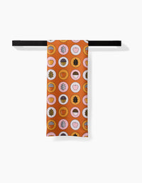 The Apples Acorns tea towel by Megan Roy features an orange, quick-drying fabric with acorn, apple, and leaf circle patterns in brown, pink, and cream, displayed on a black rod against a white background.