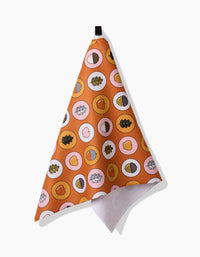 The Apples Acorns kitchen towel by Megan Roy features a vibrant orange background with circles of hearts, leaves, and acorns. Made from recycled material, this quick-drying towel hangs on a white wall by a black loop.