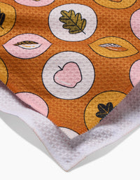 A close-up of Megan Roys Apples Acorns recycled kitchen towel shows a brown, pink, and yellow leaf and apple pattern in circles with a white, waffle-textured edge.