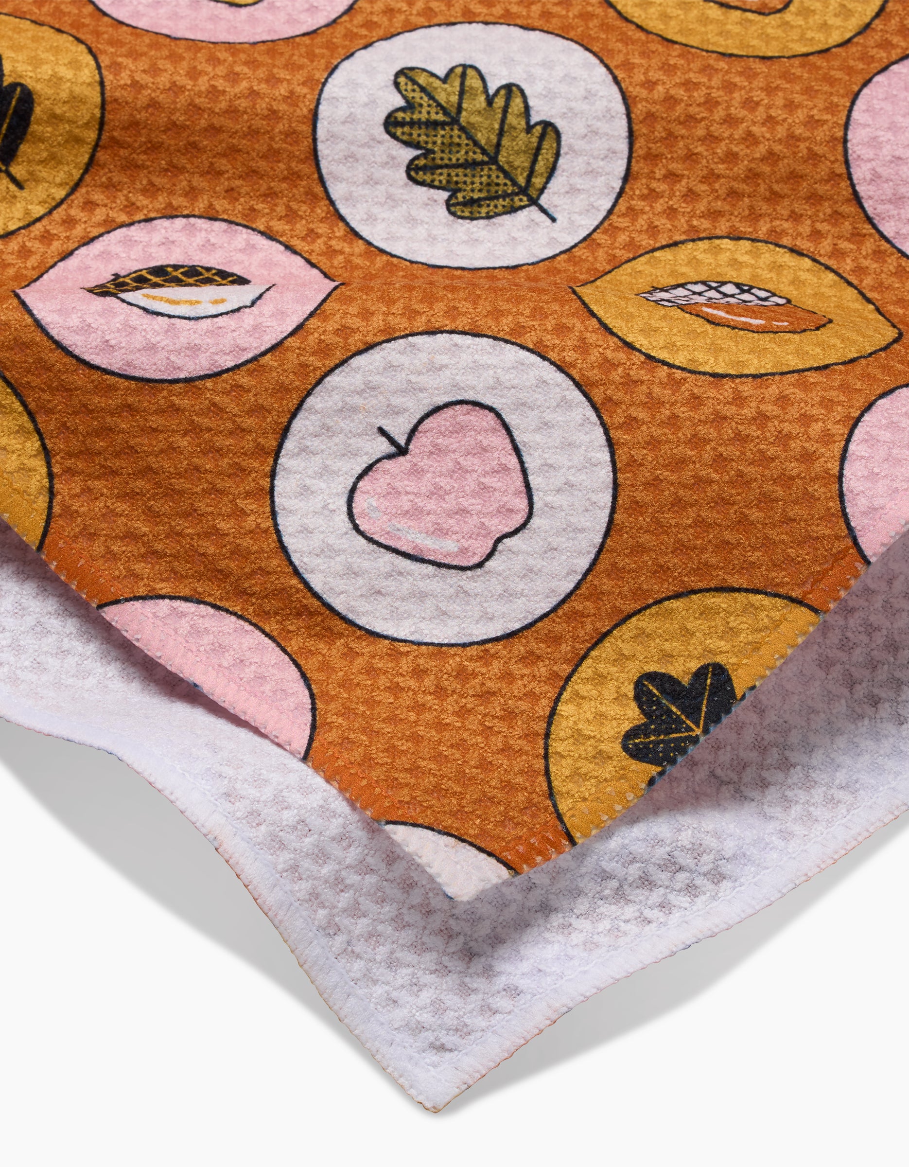 A close-up of Megan Roys Apples Acorns recycled kitchen towel shows a brown, pink, and yellow leaf and apple pattern in circles with a white, waffle-textured edge.