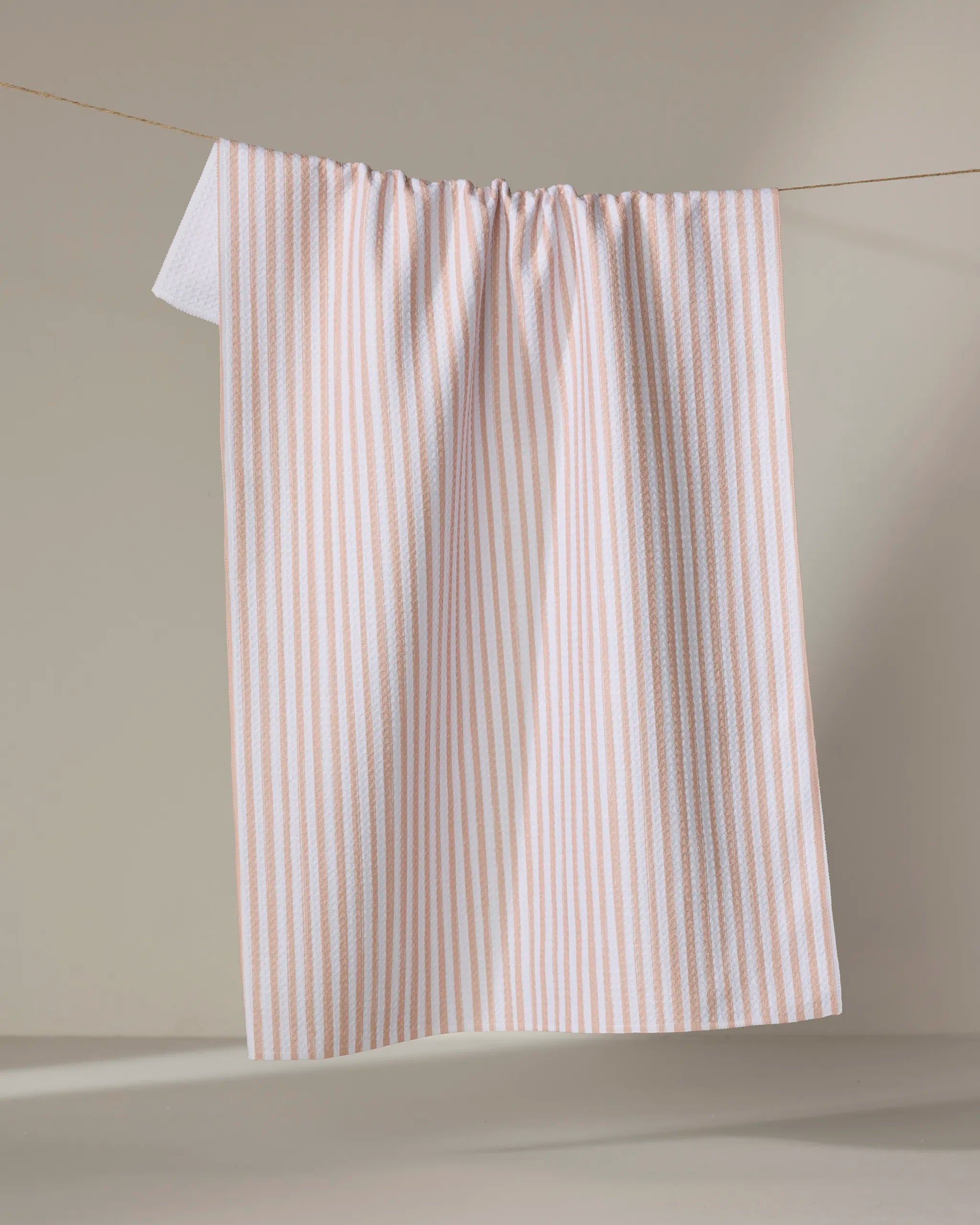 The Kailee Wright "Apricot" towel, featuring light pink and white vertical stripes and quick-drying fabric, hangs from a string against a plain beige background.