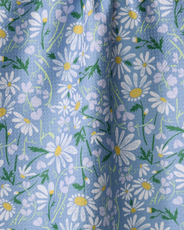 A close-up of the Elisabeth Wing "April" tea towel, ultra-absorbent and set against a blue background, showcasing white daisies with yellow centers, small lavender flowers, plus green leaves and stems in its floral design.