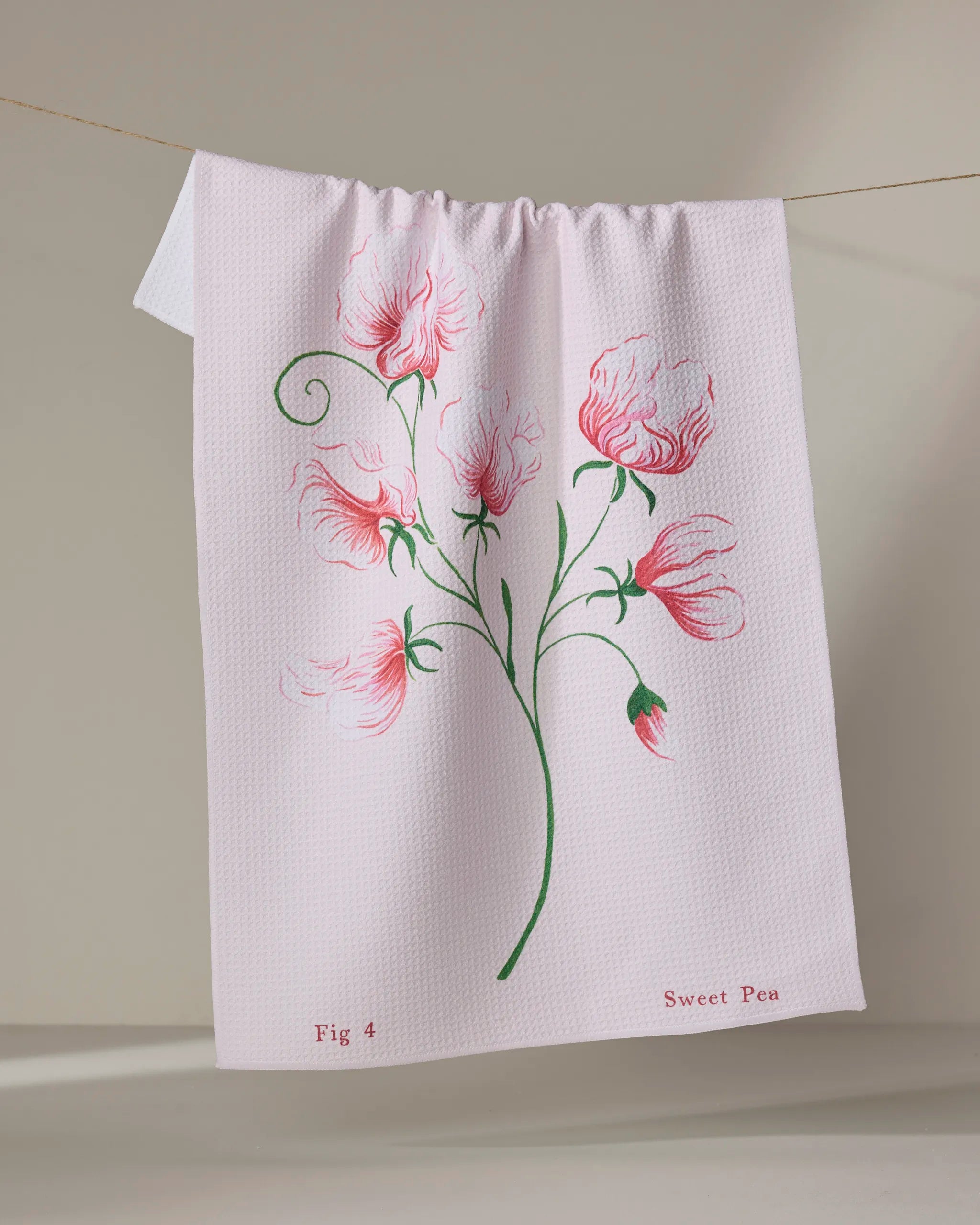 The April Sweet Pea tea towel by Linda Fox is made from ultra-absorbent recycled material, with pink sweet pea flowers and green stems. As it hangs, "Fig 4" and "Sweet Pea" appear in red at the bottom corners.