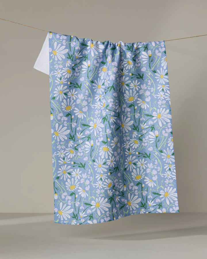The April tea towel by Elisabeth Wing features a white daisy and green leaf pattern on blue, hanging on a clothesline with a neutral background. Made from recycled materials, it’s an eco-friendly and charming addition to any home.