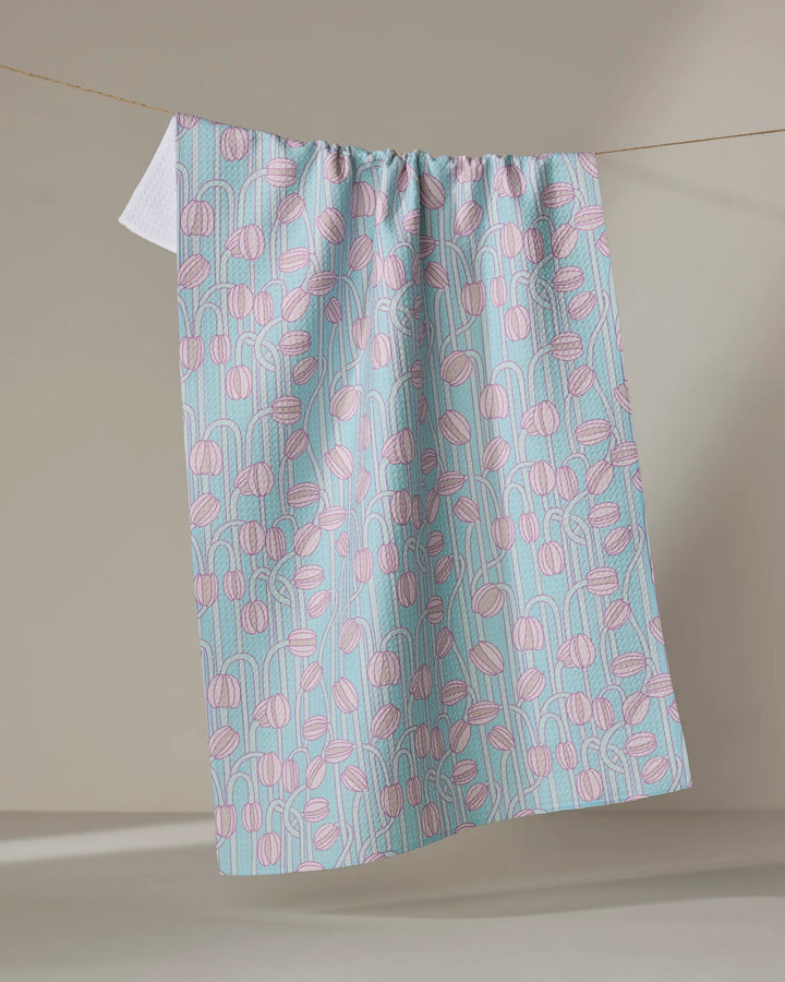 The Aqua Poppy Pods towel by Katie O'Shea, featuring pastel pink abstract flowers and blue vertical lines, hangs on a clothesline against a neutral background.