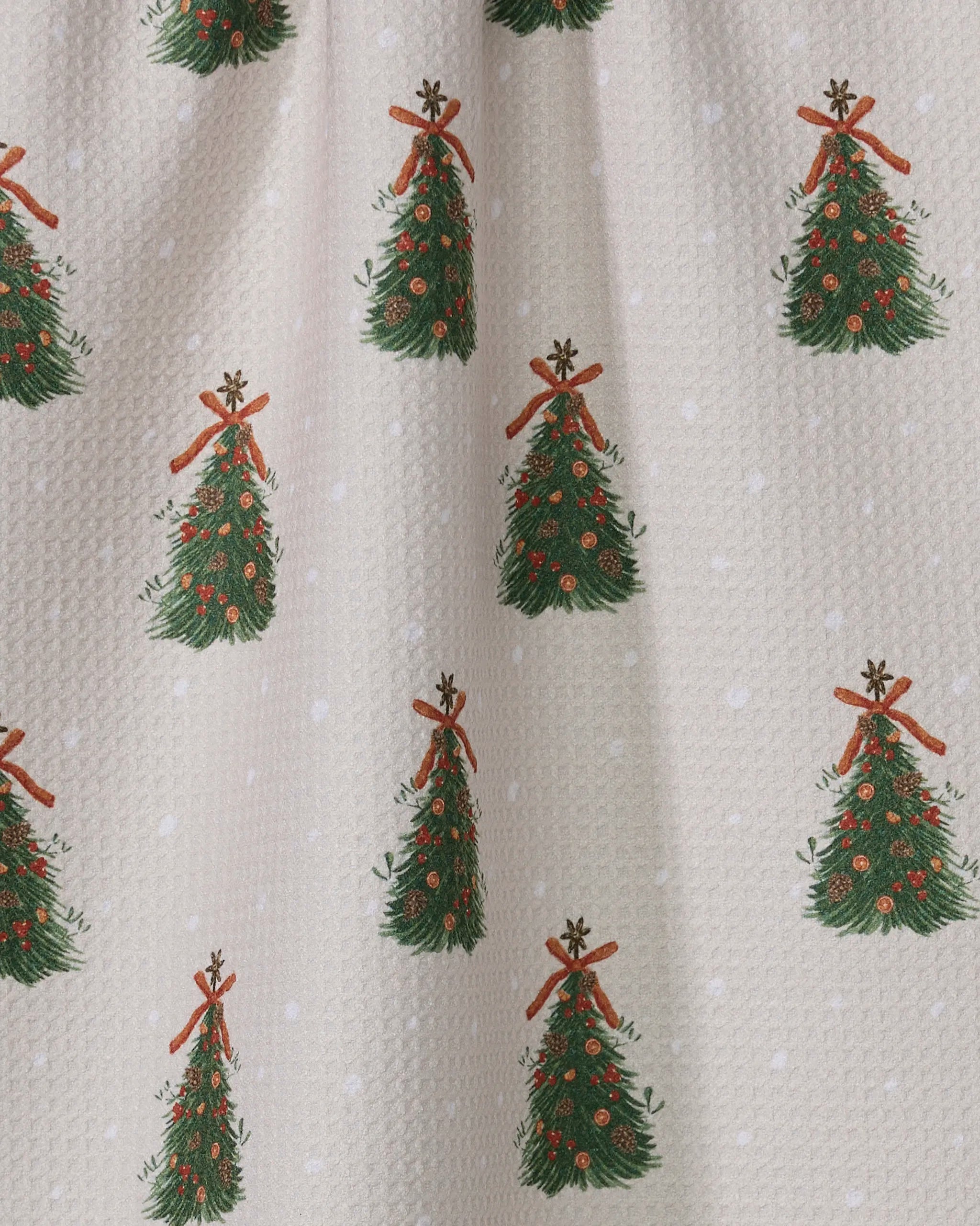 The Arbolitos tea towel by Geometry is beige, made from recycled materials, and features green Christmas trees with red ornaments, brown stick stars, and white snow-like dots.