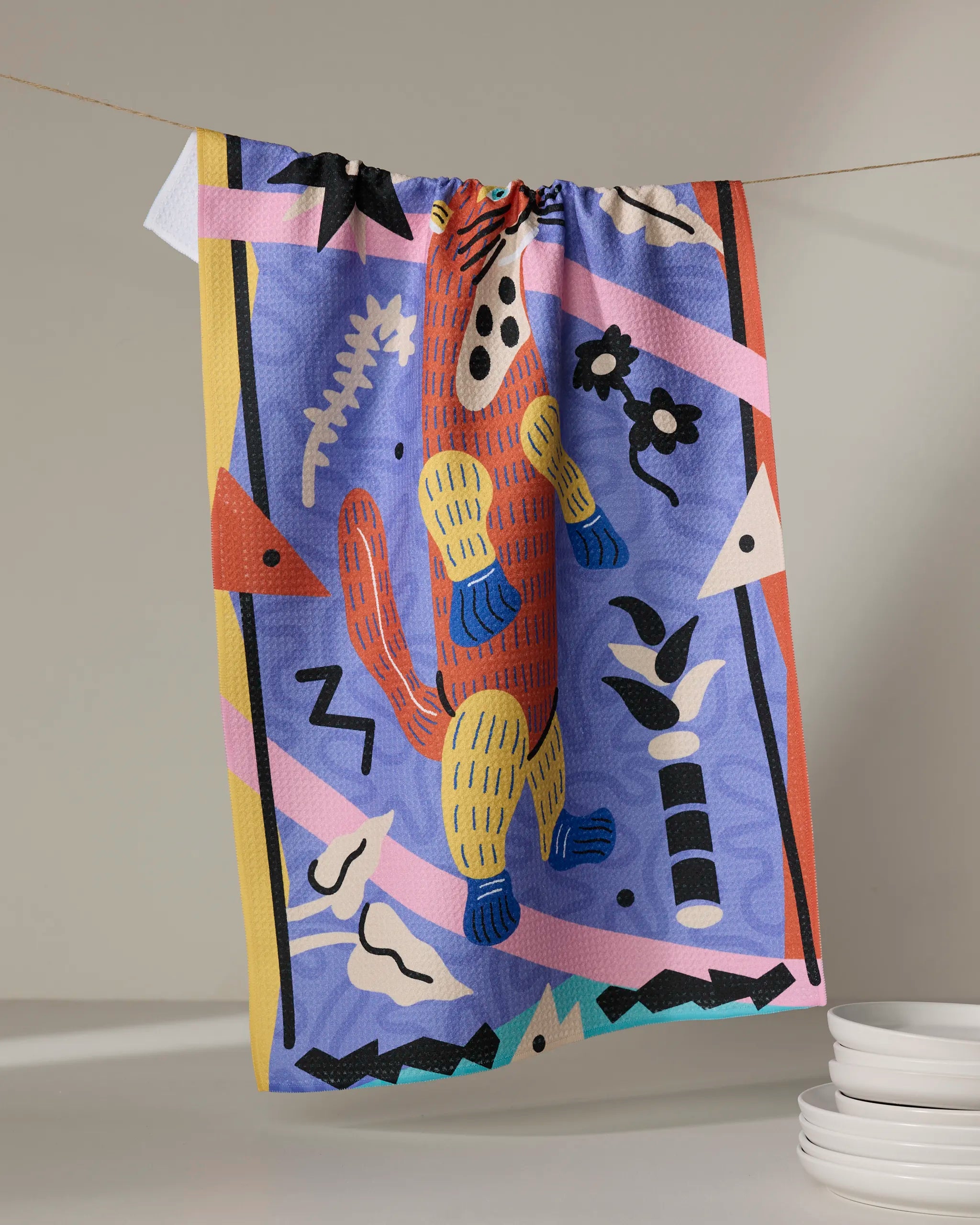 The Ariranha tea towel by Camila Leão, made from recycled materials, showcases a vibrant abstract design with a playful animal and bold patterns, hanging above white plates.