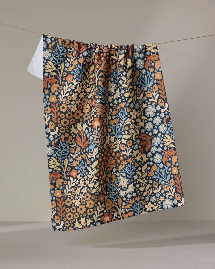 The Arthur recycled kitchen towel by Danelys Sidron, featuring a blue, orange, and cream floral pattern, hangs on a clothesline against a neutral background.