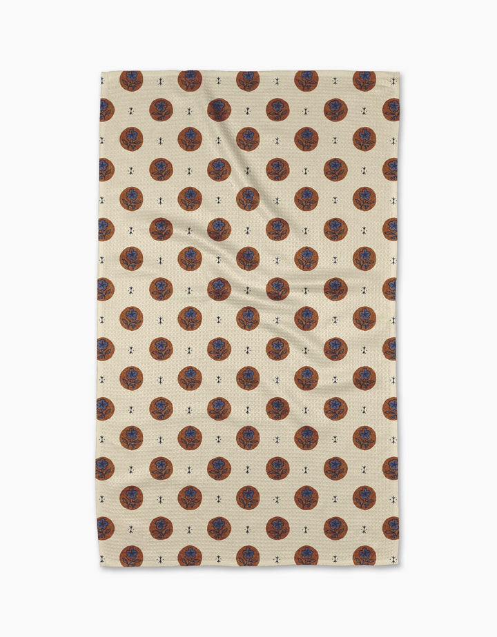 A beige Geometry Artisan Floral Tea Towel with a repeating pattern of red and blue circular floral designs and small black accents, displayed flat against a white background.