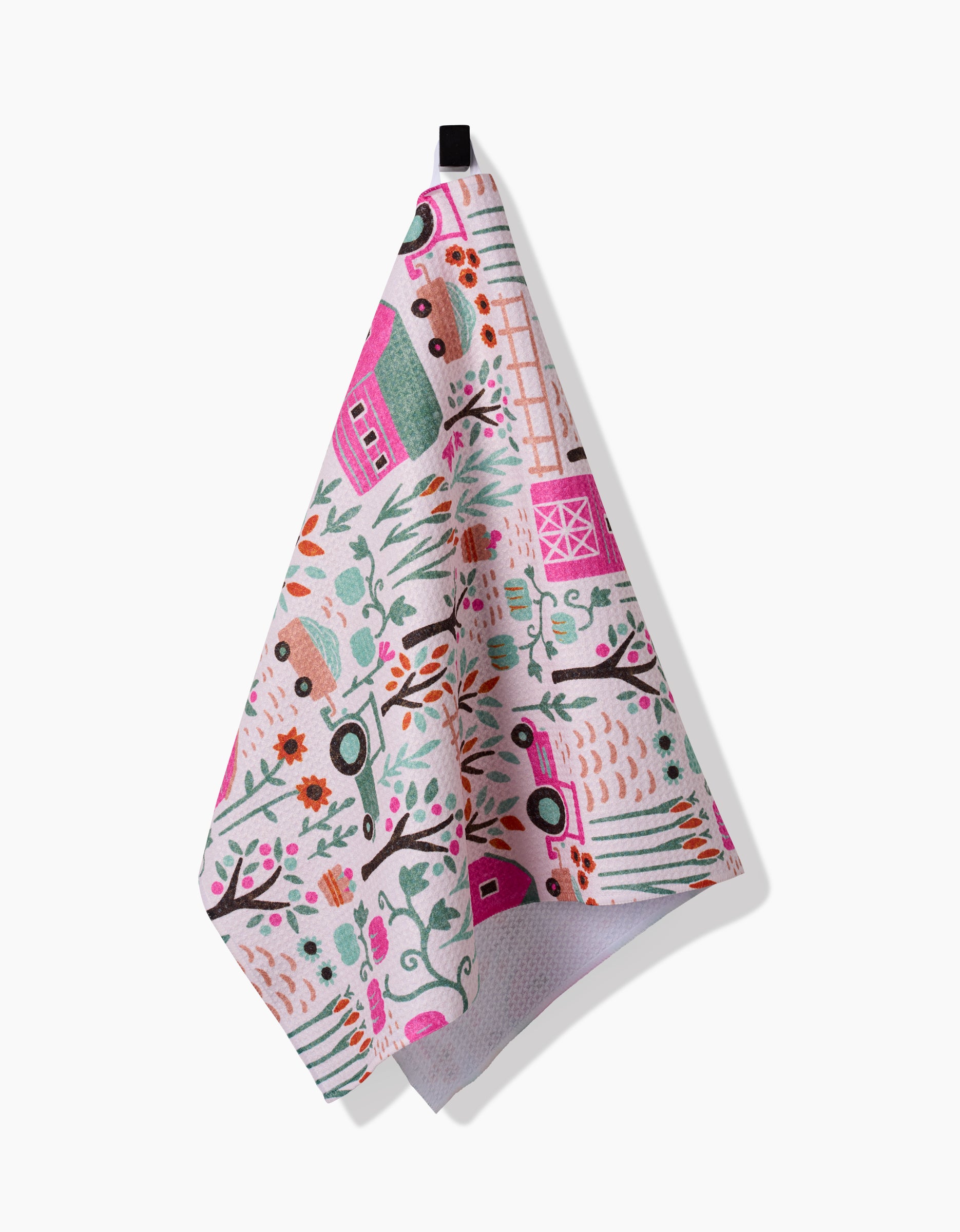 The Amy MacCready Asher tea towel features a colorful countryside pattern with barns, tractors, trees, and plants in pink, green, and orange shades. It’s ultra-absorbent and hangs from a black loop against a white background.