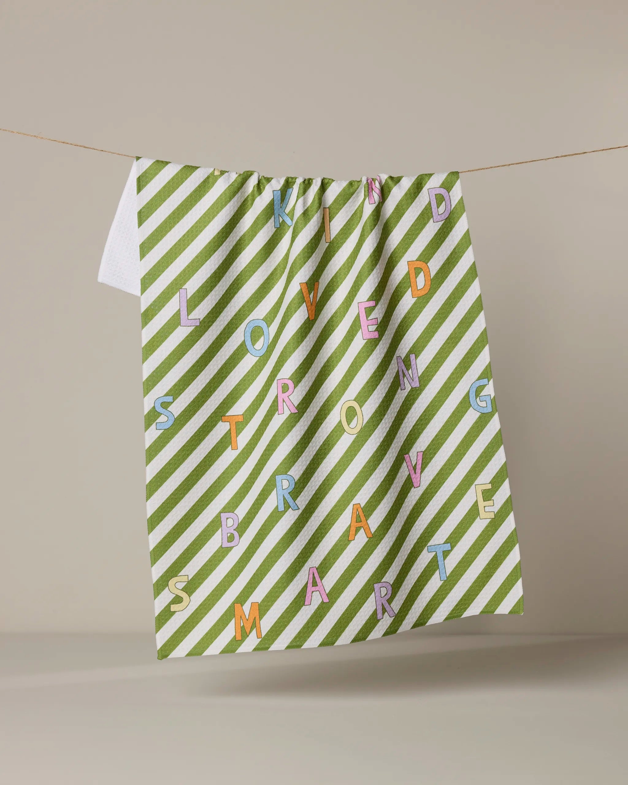 A Geometry Attributes recycled kitchen towel with green and white diagonal stripes hangs on a line. The towel features colorful lettering spelling out KIND, LOVED, STRONG, BRAVE, and SMART.
