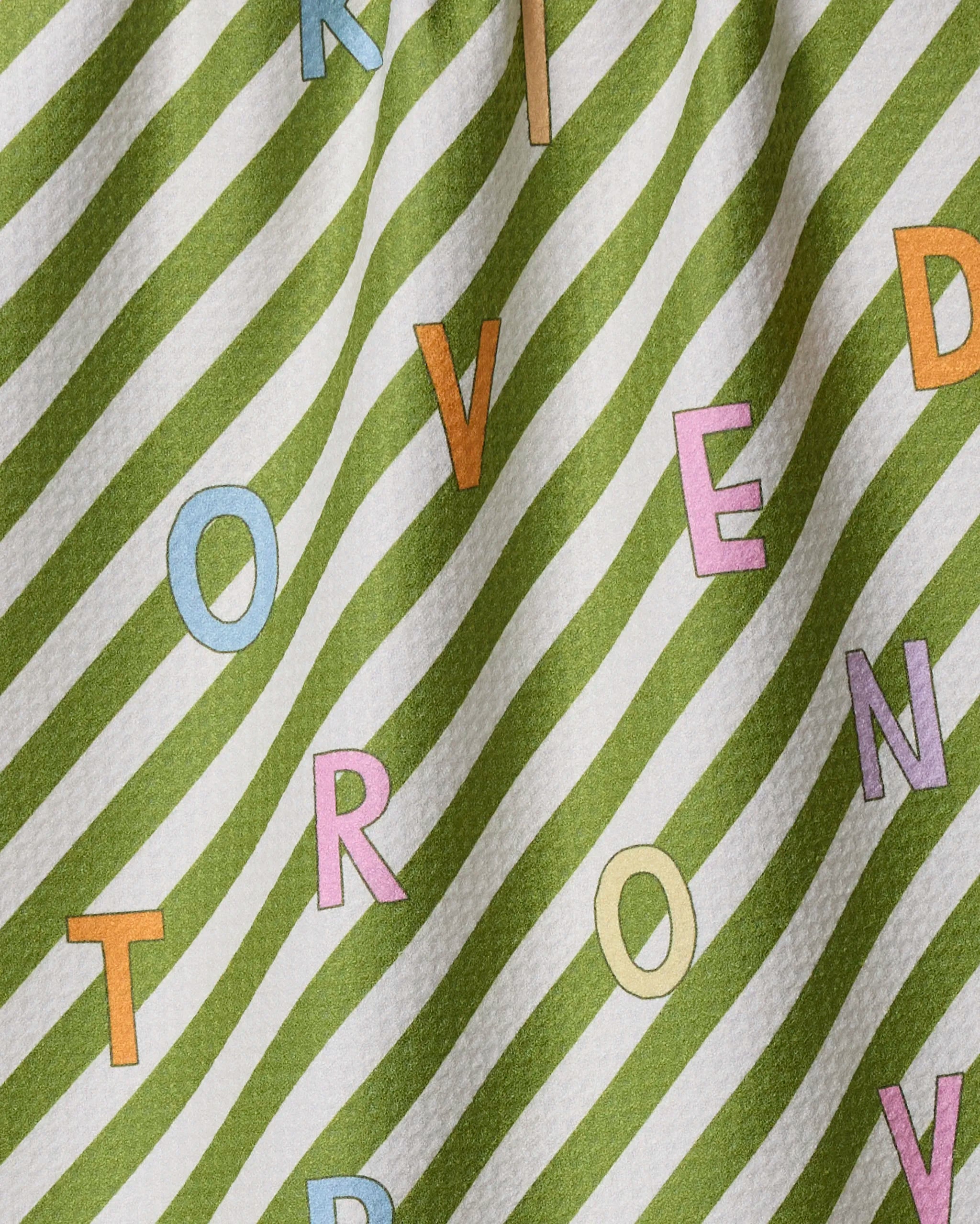 The Geometry Attributes dish towel features green and white diagonal stripes with large, colorful random letters (O, V, E, D, T, R, N, K) scattered across its absorbent fabric and a slightly wavy texture.