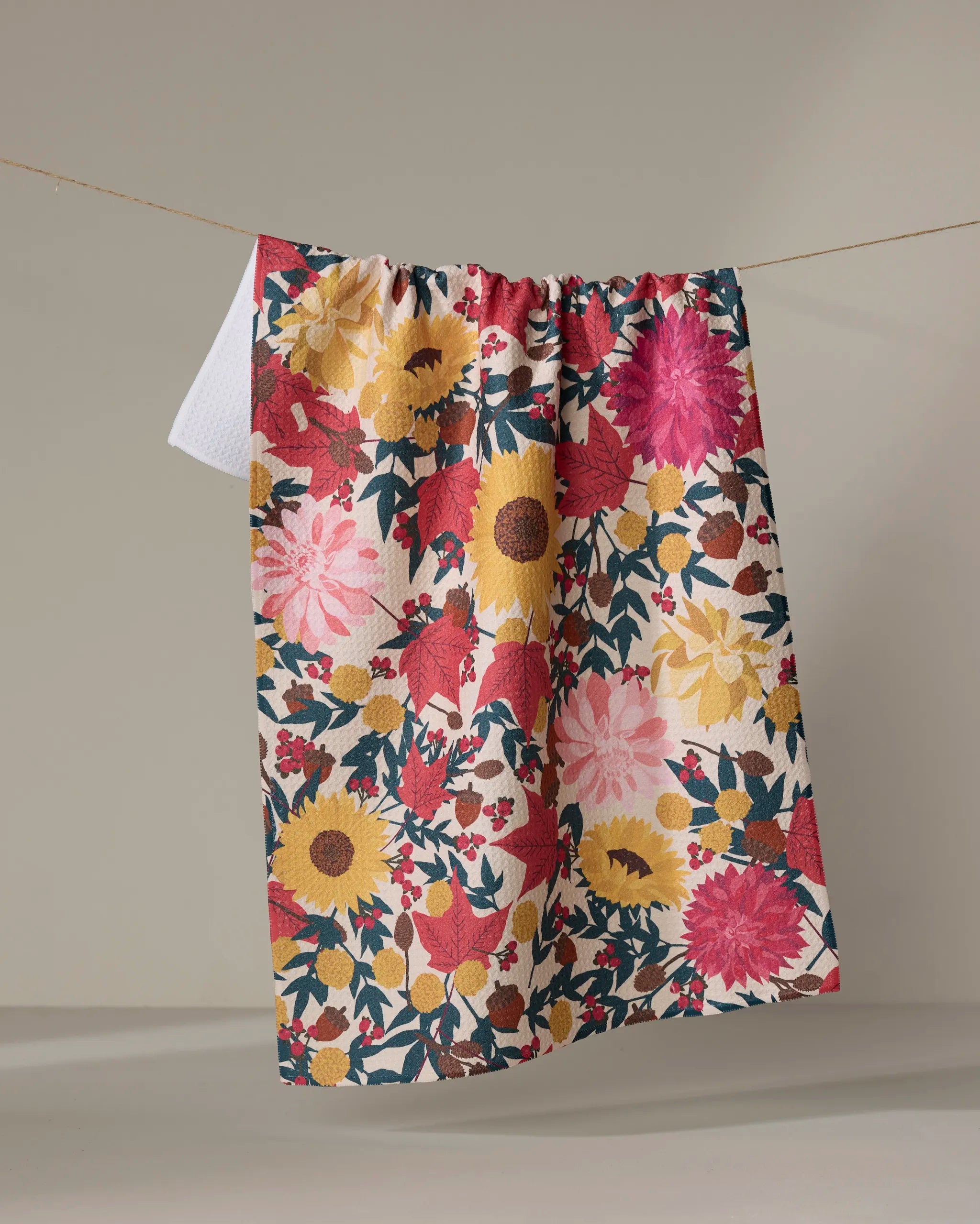 The "Autumn Blooms" tea towel by Rebecca Bobko, featuring a vibrant floral design in red, pink, yellow, and green, hangs from a thin string against a plain, neutral background.