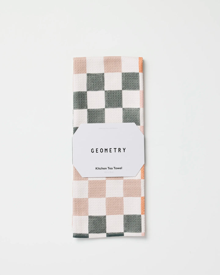 The GEOMETRY Autumn Checkers kitchen tea towel, featuring a white, gray, and light beige checkered pattern, is folded and wrapped with a white label reading "GEOMETRY Kitchen Tea Towel" on a white background.