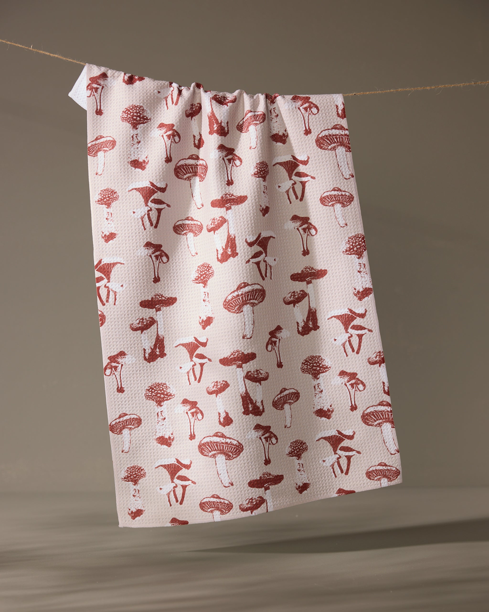 The Geometry Autumn Mushrooms tea towel, made from recycled materials and featuring a red mushroom and animal pattern, hangs on a string against a neutral background.
