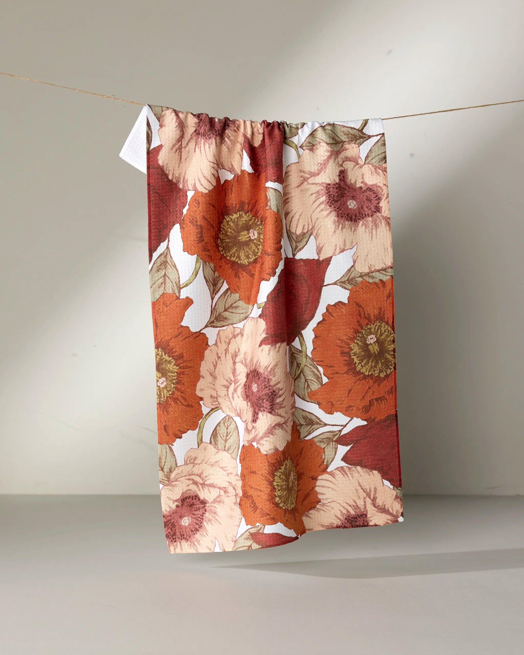 The "Autumn Petals" tea towel by Ashley Reeves, featuring a bold floral pattern in red, orange, and cream, hangs from a string against a neutral background.