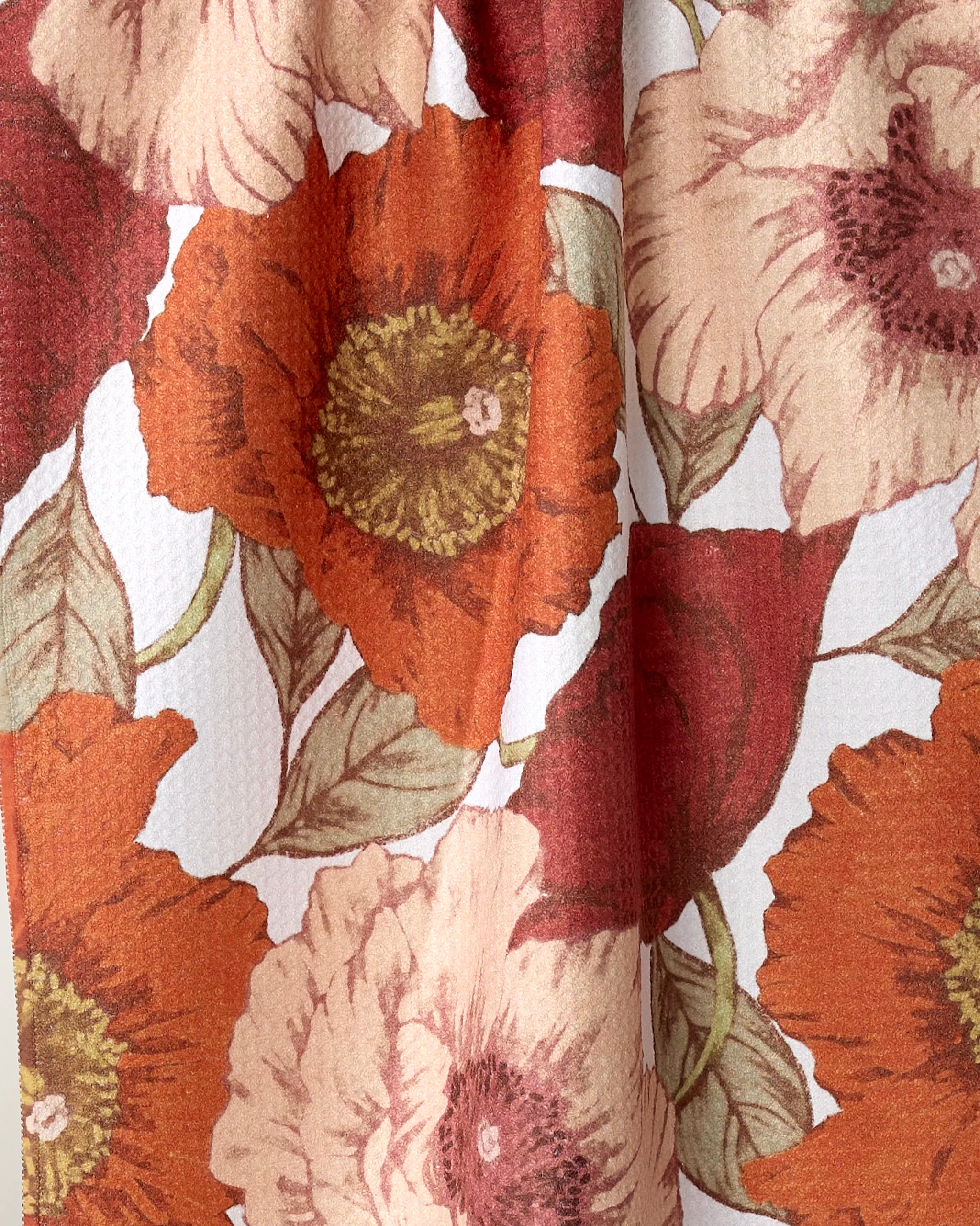 A close-up of the "Autumn Petals" tea towel by Ashley Reeves, made from recycled materials. It showcases large red, orange, and beige flowers with green leaves on a white background and has a slightly visible texture.