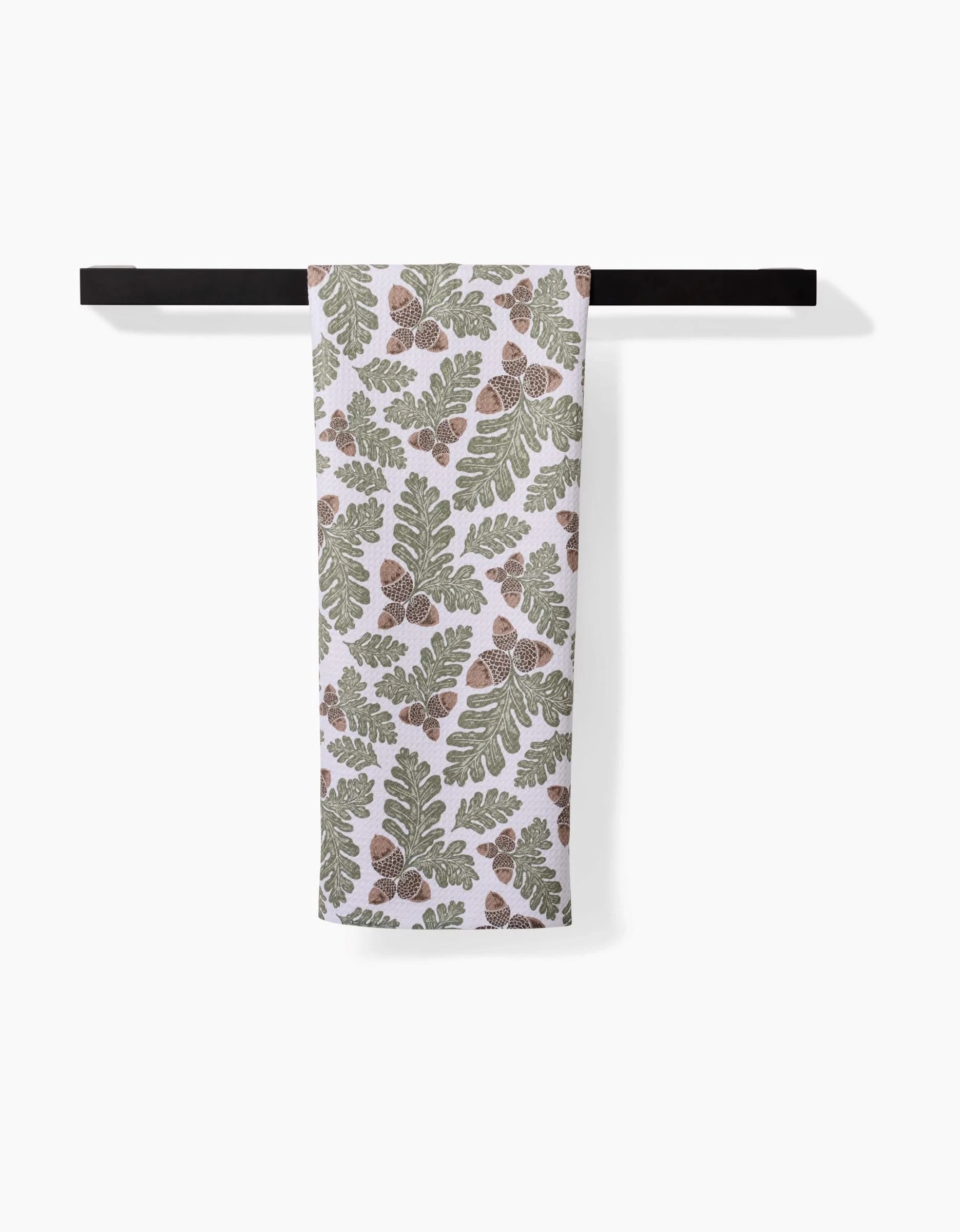 The Danelys Sidron Autumn Pines towel, featuring green oak leaves and brown acorns on white ultra-absorbent Geoweave fabric, hangs on a black horizontal towel bar against a plain white background.