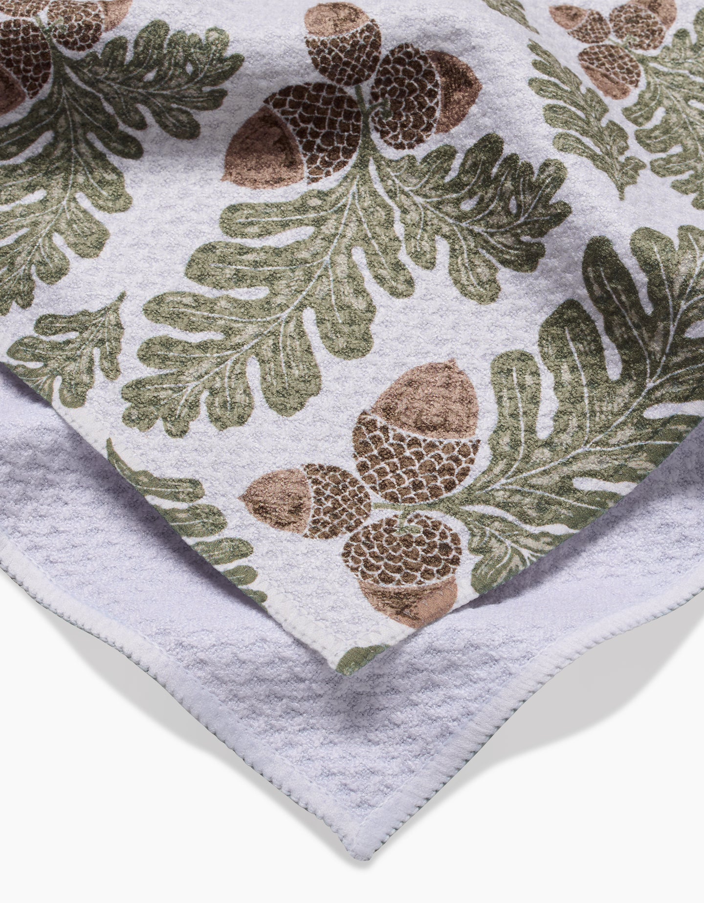 The Autumn Pines towel by Danelys Sidron features ultra-absorbent Geoweave fabric with a green oak leaf and brown acorn pattern. The image shows it folded, revealing part of the plain underside.
