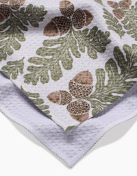The Autumn Pines towel by Danelys Sidron features ultra-absorbent Geoweave fabric with a green oak leaf and brown acorn pattern. The image shows it folded, revealing part of the plain underside.