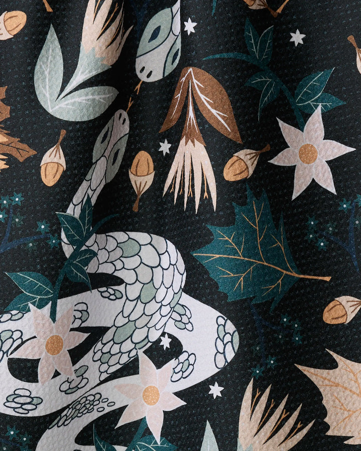 Julianne Haness's "Autumn Snakes" tea towel features a white snake with flowers, acorns, and leaves on a dark background with whimsical stars. Made from recycled materials, this ultra-absorbent kitchen towel is both stylish and eco-friendly.