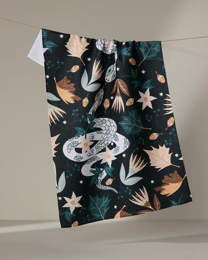 The "Autumn Snakes" kitchen towel by Julianne Haness is ultra-absorbent, patterned with a white snake amid earthy-toned leaves, flowers, and stars, displayed hanging against a neutral background.