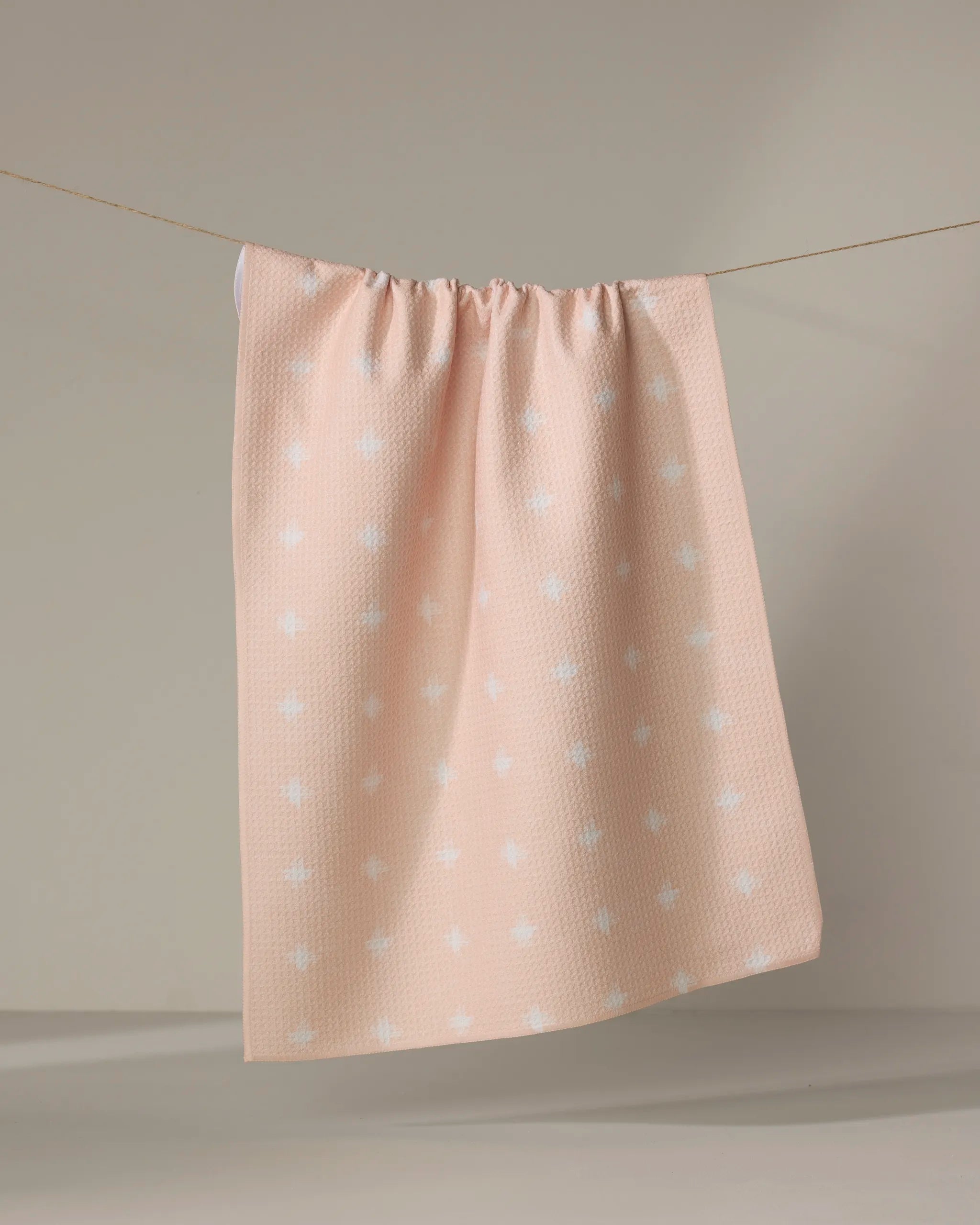 The Aveline tea towel by Amy MacCready, featuring a light pink textured fabric with small white cross patterns, hangs from a clothesline against a neutral background.