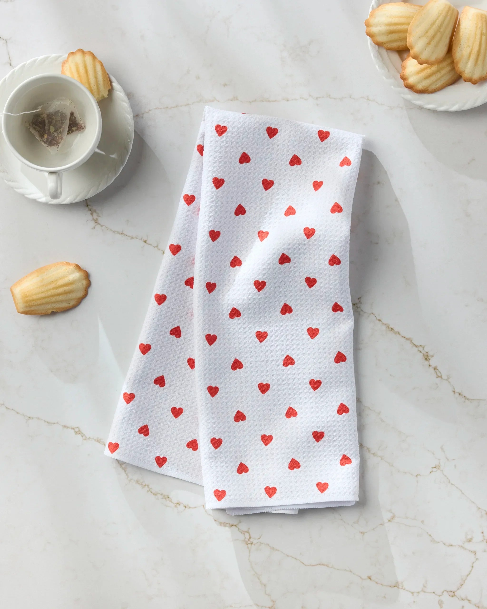 The Geometry "Baby Hearts" tea towel, made from ultra-absorbent recycled materials and featuring red heart patterns, is displayed on a marble surface beside a teacup, saucer, and madeleine cookies.