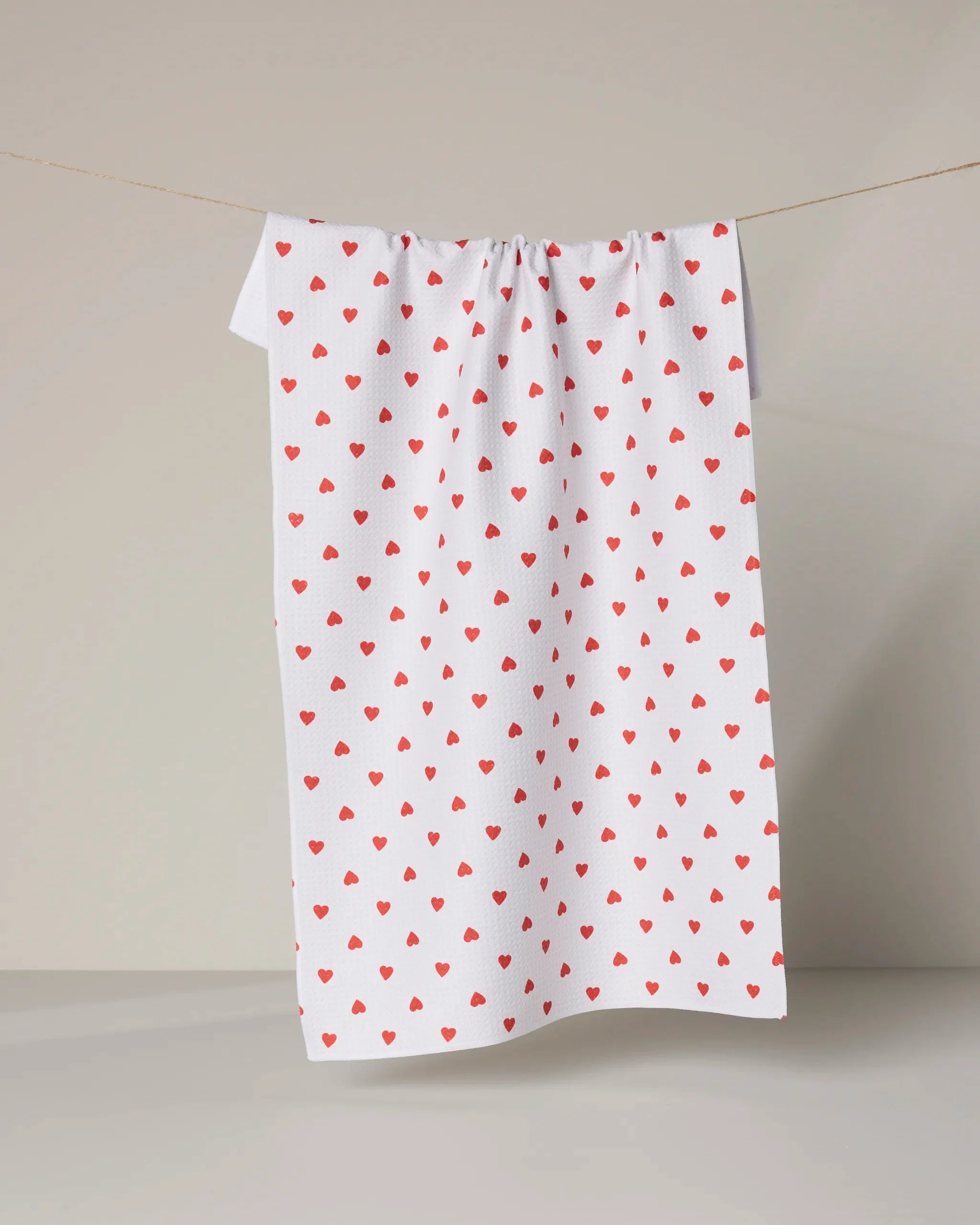 The Geometry "Baby Hearts" towel, ultra-absorbent and decorated with small red hearts on white, hangs on a clothesline against a plain light background.