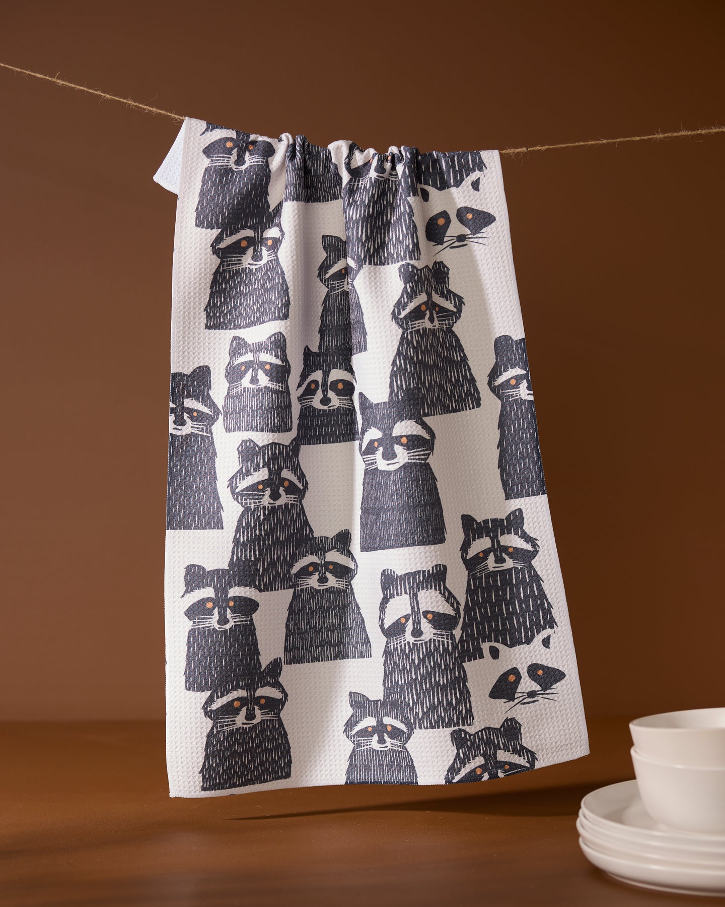 The Geometry Banditbunch white tea towel, featuring a black and grey raccoon pattern, hangs on a string against a brown background beside a stack of white dishes.