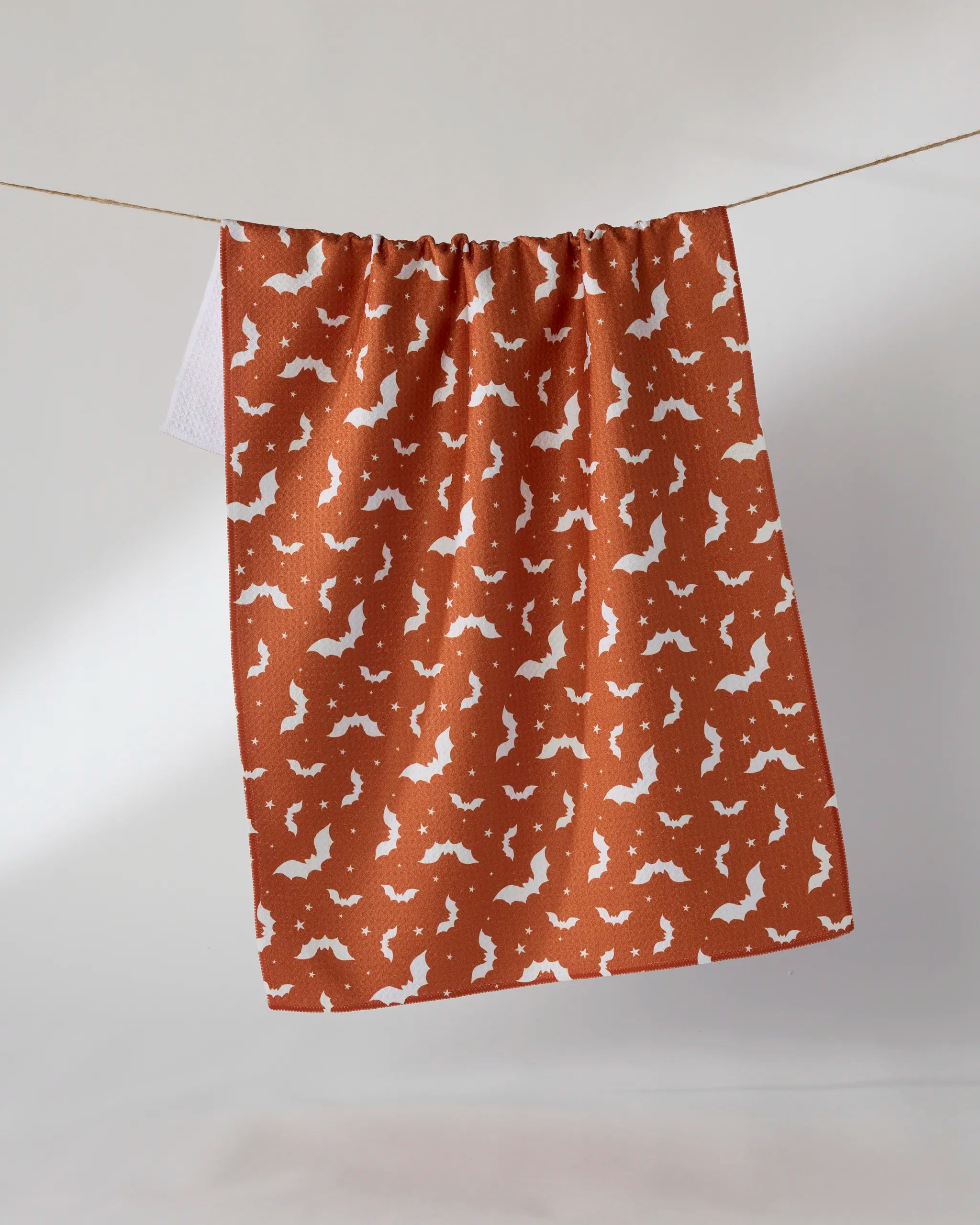 The Geometry "Batty Night" tea towel, featuring a white bat pattern on burnt orange, hangs on a clothesline against a plain light background.