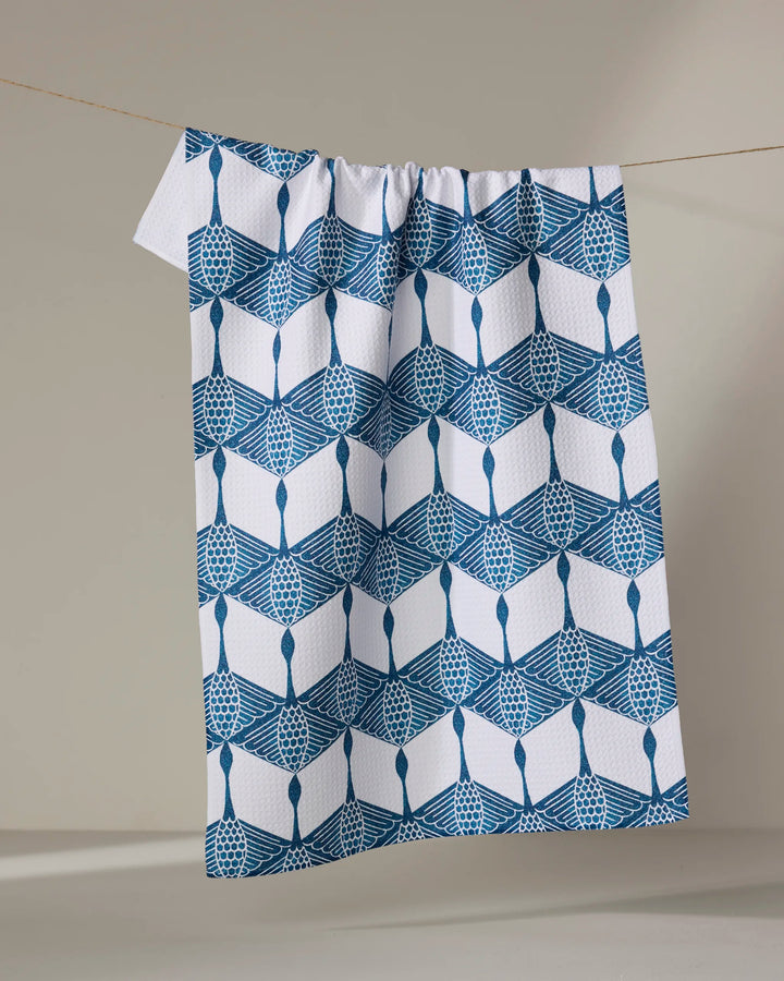 The BB Cubed kitchen towel by Renee Fly, made from recycled materials, features a blue geometric and lattice pattern on white and hangs from a clothesline against a neutral background.