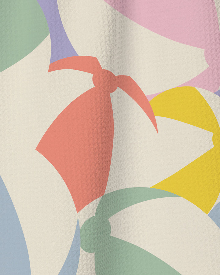 The Geometry "Beach Ball" tea towel features an abstract design with overlapping pastel shapes—green, pink, purple, yellow, and red—on a cream textured background, including forms that evoke stylized cherries and leaves.