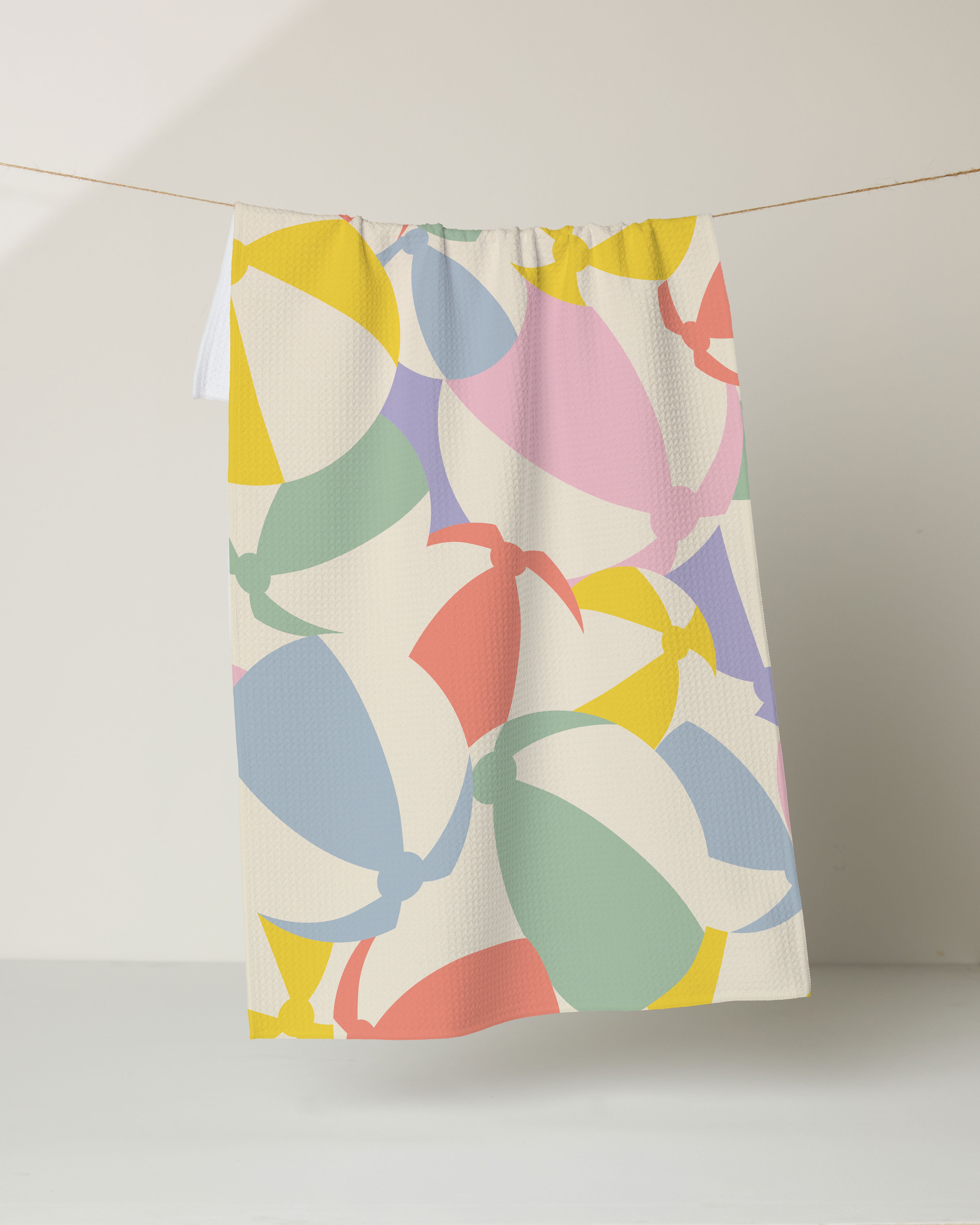 The Geometry Beach Ball tea towel features overlapping beach balls in pastel yellow, green, red, purple, and blue, hanging from a clothesline against a white background.