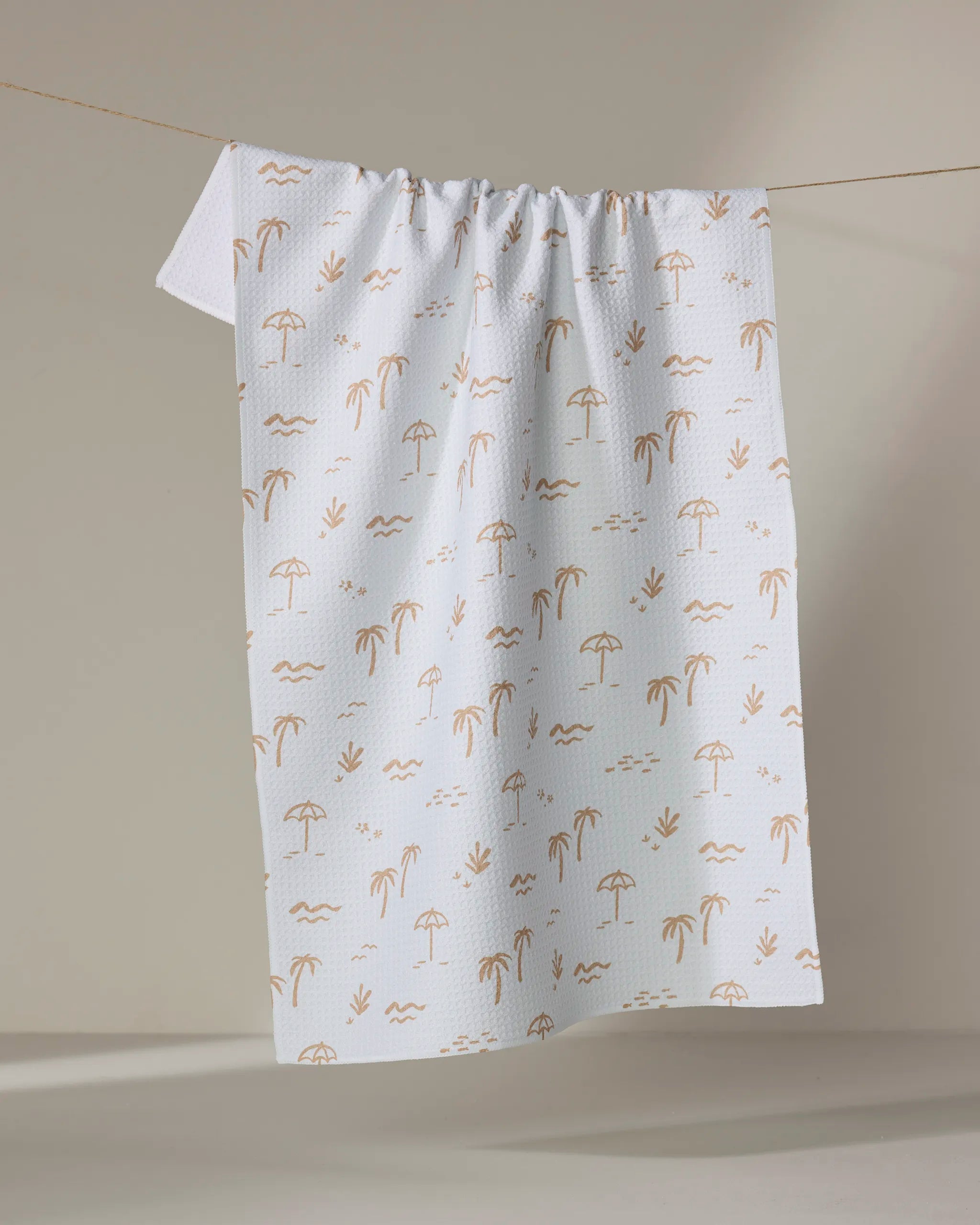 The "Beach" tea towel by Jen B. Peters is made from recycled materials and features a light brown beach pattern with palm trees, umbrellas, waves, and seagulls, shown hanging on a string against a neutral background.