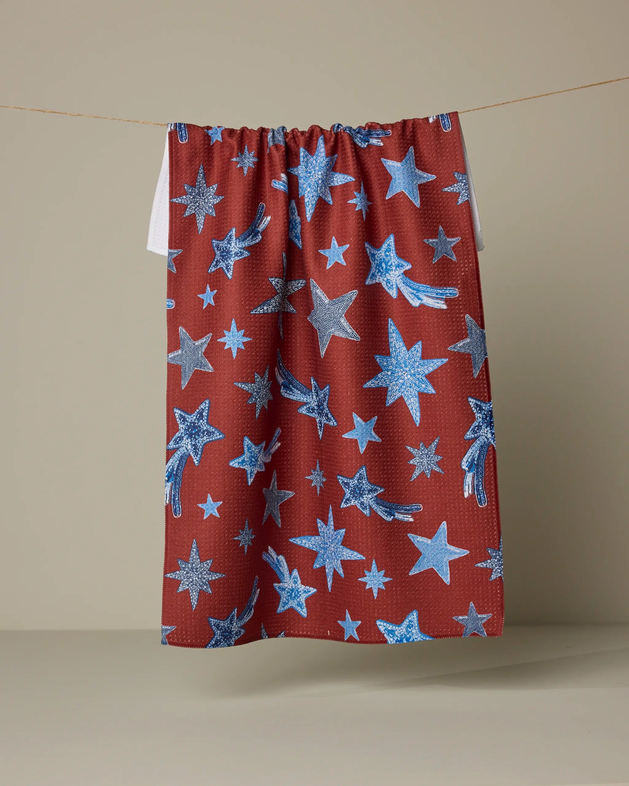 The Geometry "Beaded Stars" tea towel, featuring a blue star and shooting star pattern on red, made from recycled materials, hangs against a beige background.