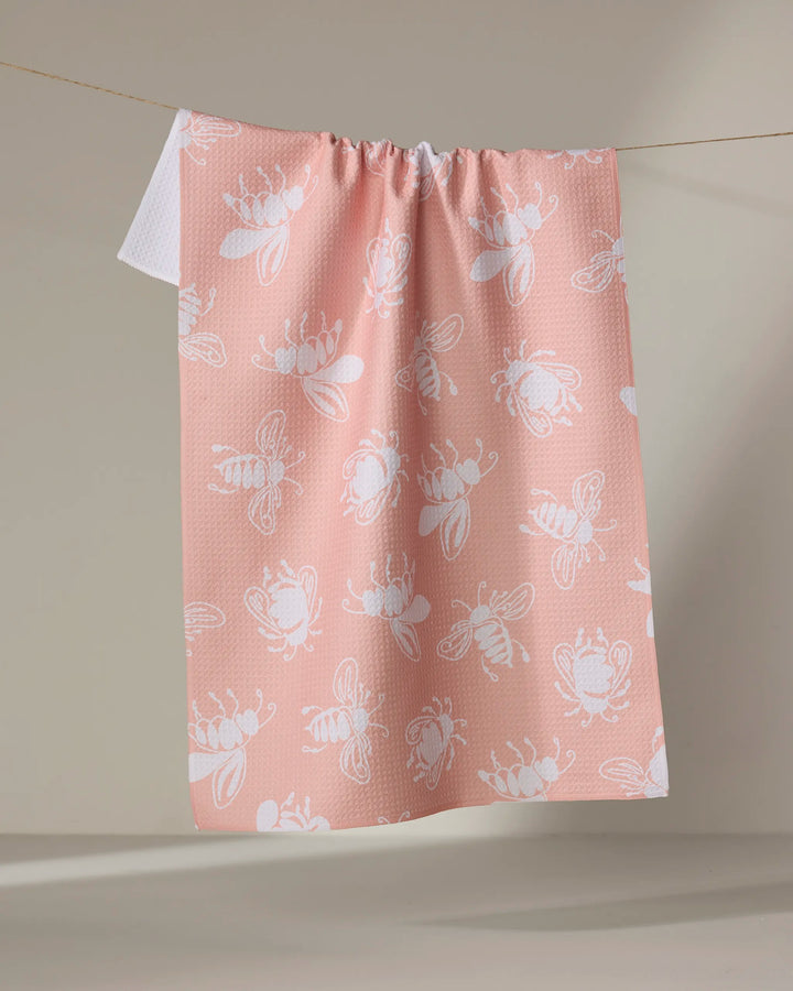 The Sara Mari Beelieve pink tea towel, featuring a white bee pattern and crafted from recycled materials, hangs on a clothesline against a plain beige background.