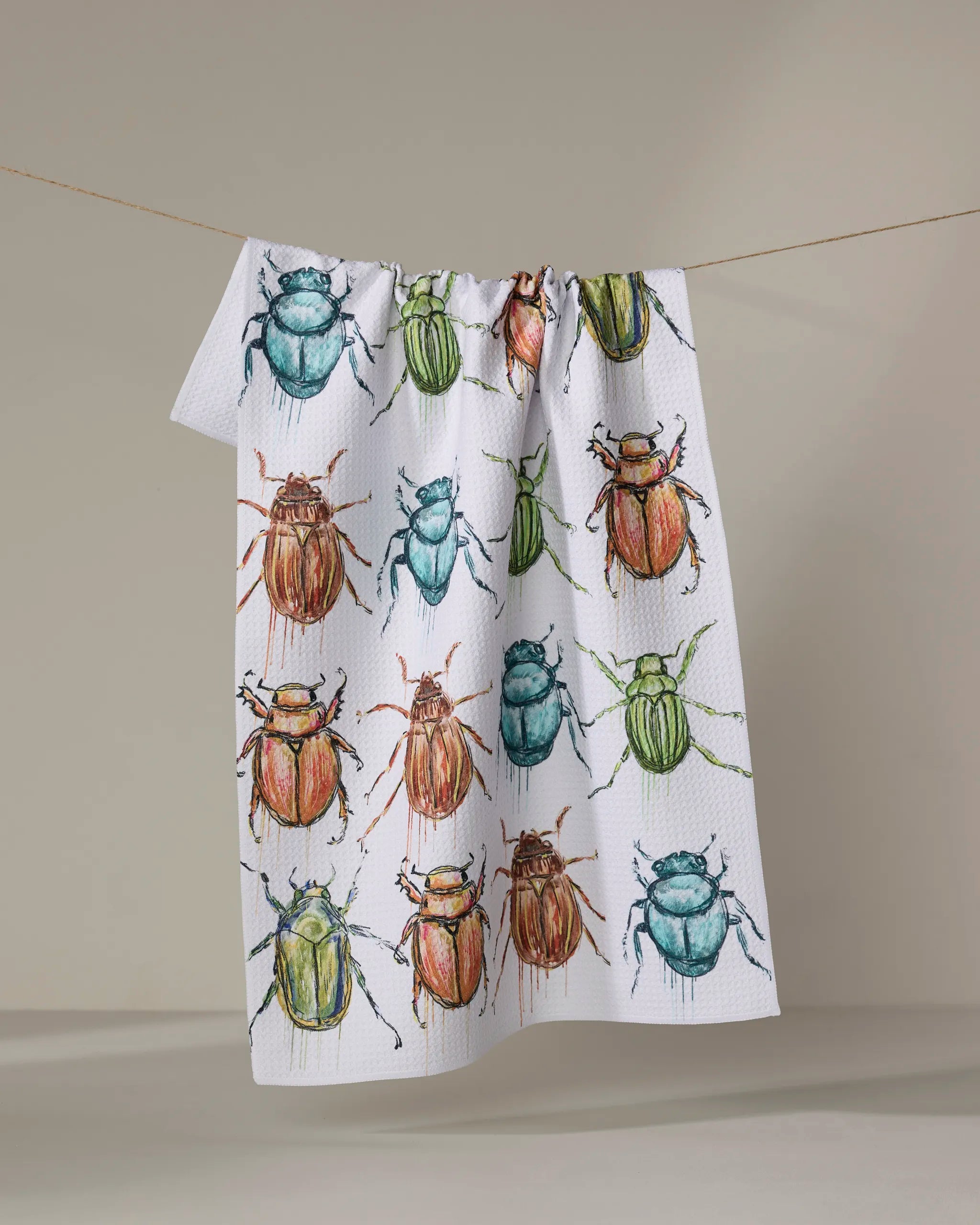 The Taryn Brennan "Beetles" tea towel, ultra-absorbent and white with vibrant red, green, and blue beetle illustrations, hangs on a line against a neutral background.