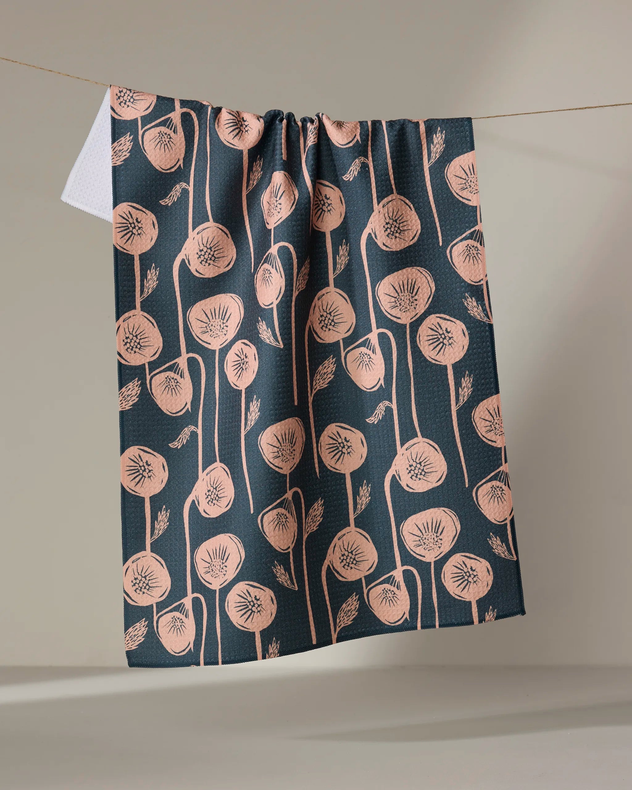 The "Belgian Poppies" tea towel by Elisabeth Wing showcases a dark blue, quick-drying fabric with a peach botanical print of stylized, dandelion-like flowers, elegantly draped over a line against a neutral backdrop.