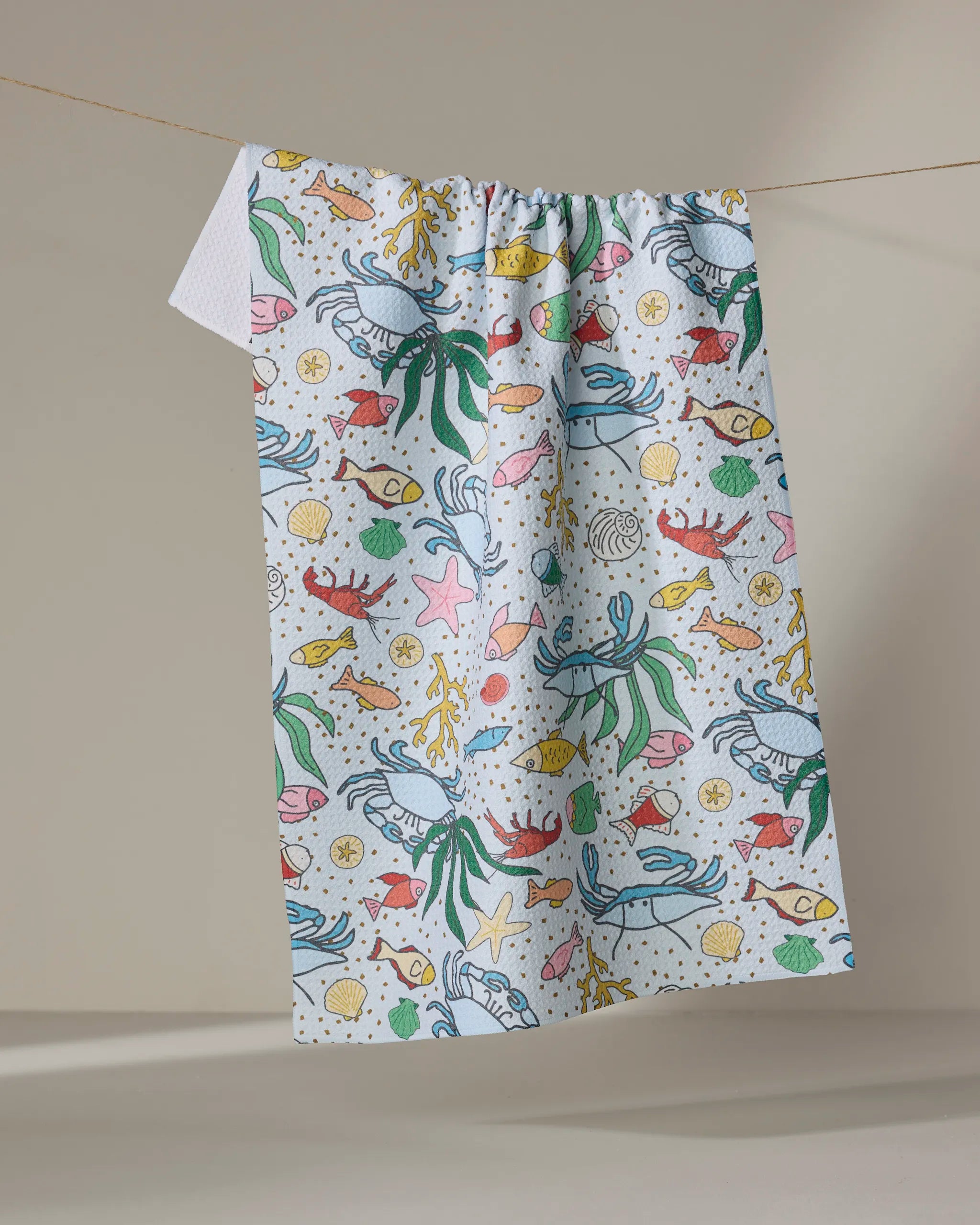 The "Beneath the Waves" quick-drying white tea towel by Mindfulnice, featuring colorful cartoon ocean creatures, hangs on a clothesline against a neutral beige background.