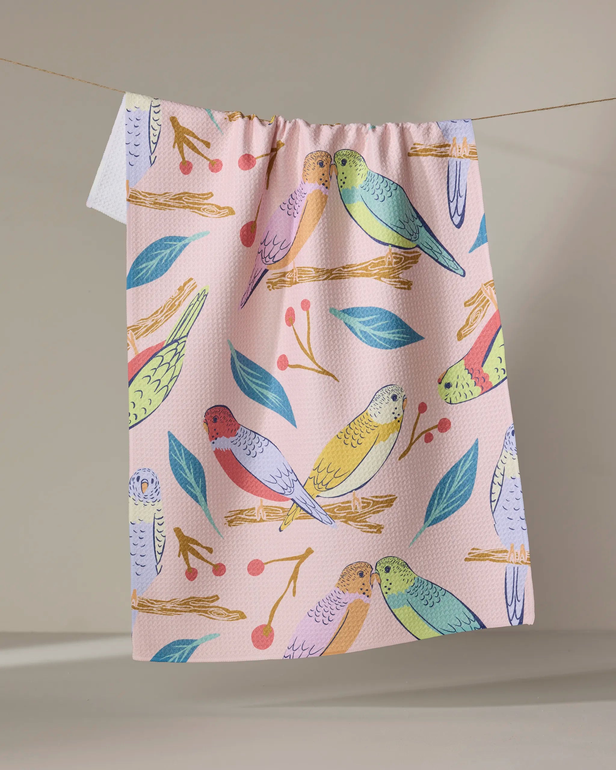 The "Berries and Budgies" tea towel by Elisabeth Wing, crafted from recycled materials, features colorful budgie birds, green leaves, and red berries on pink fabric, hanging on a string against a neutral background.