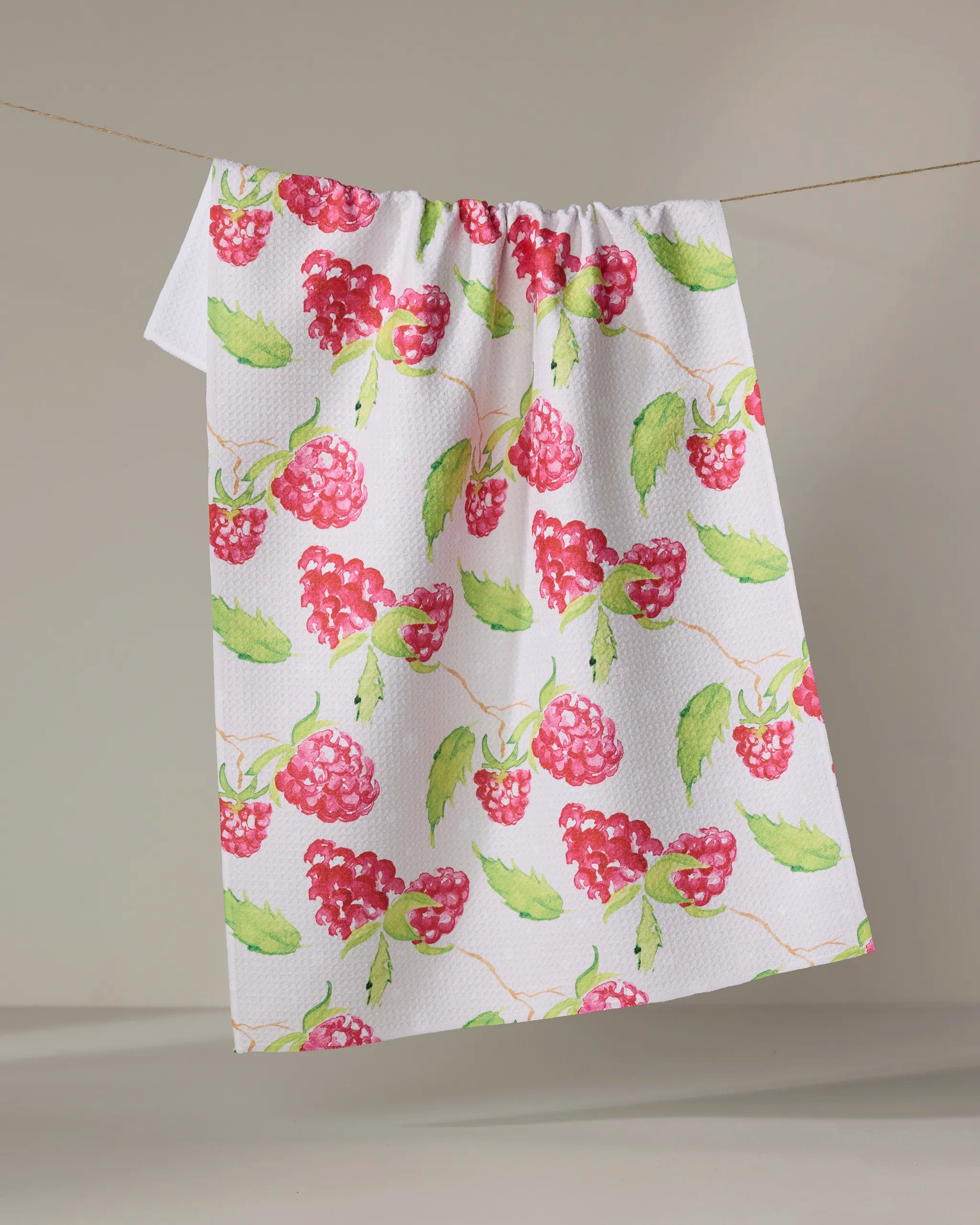 Elena Fay's Berry Bright kitchen towel, made from recycled material and featuring red raspberries with green leaves, hangs on a clothesline against a plain light background.