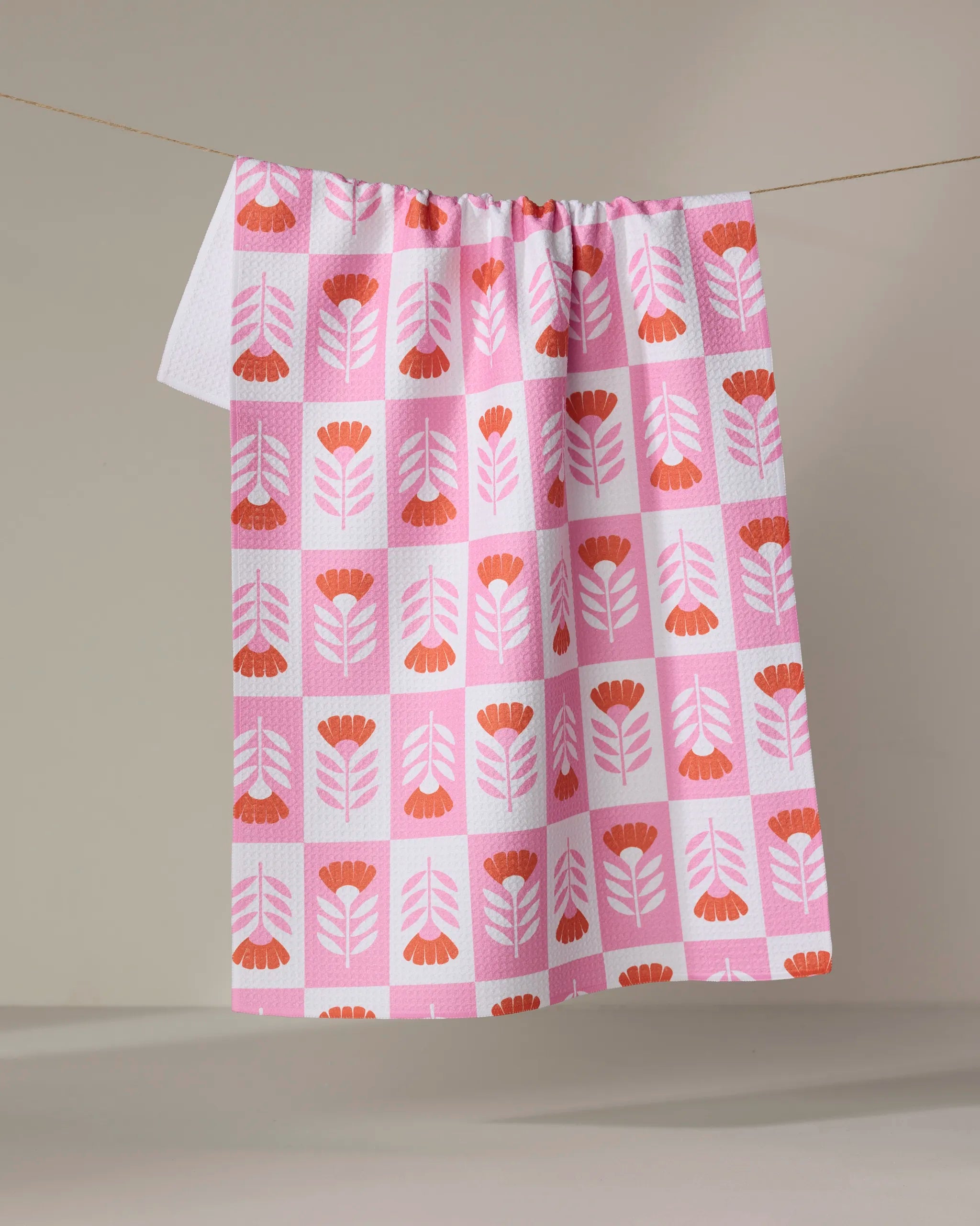 The Berry Flower Patch tea towel by Charlie Rowan Designs features a pink and white design with stylized red and orange flowers—perfect as a quick-drying kitchen towel, shown hanging on a clothesline against a neutral background.